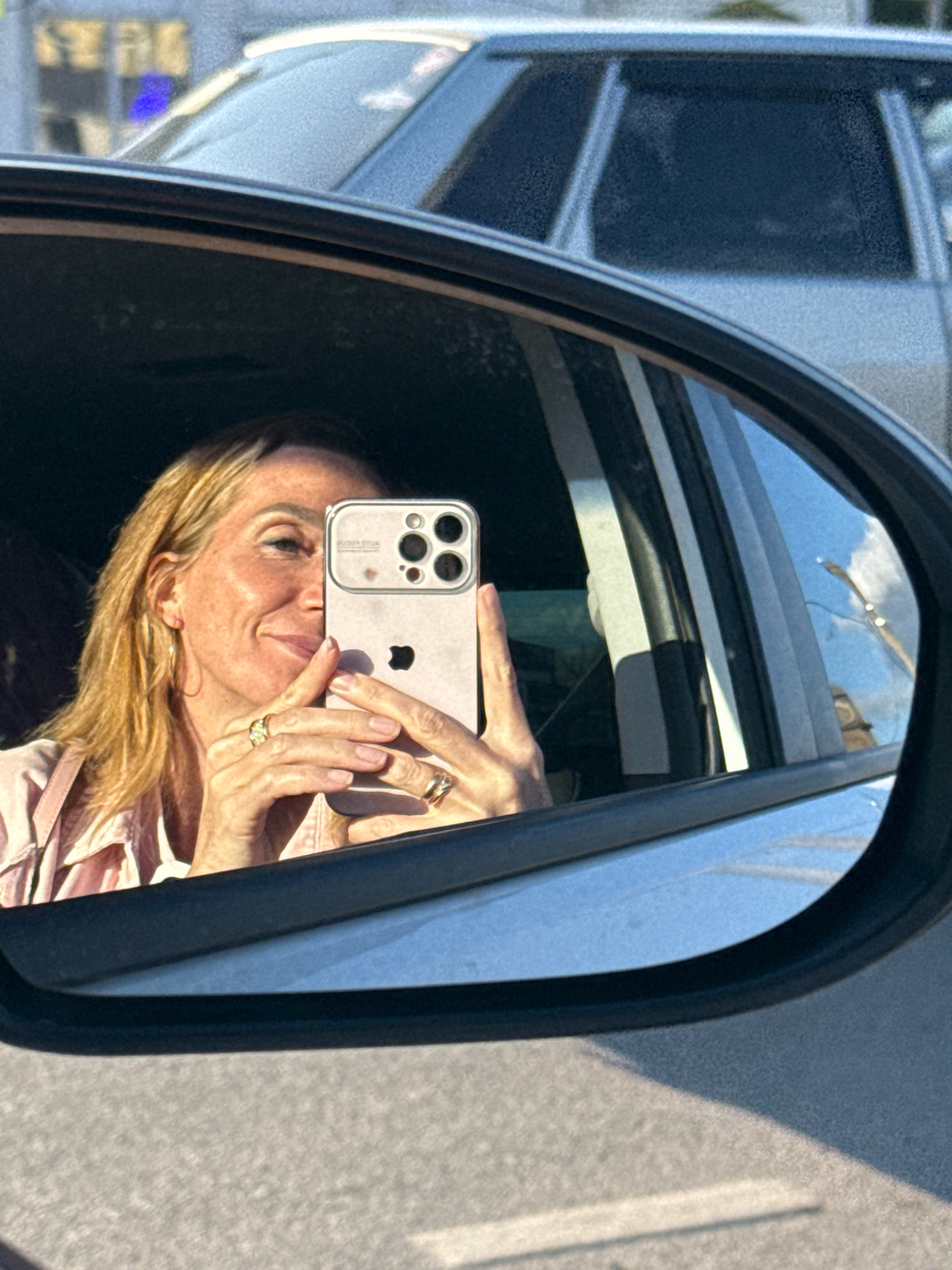 A woman is reflected in a car's side mirror, taking a selfie with her phone. She has blonde hair and is wearing a pink shirt and jewelry. The background shows cars and a blurry cityscape. The image appears to have been taken on a sunny day. The location is Torzhok, Russia.