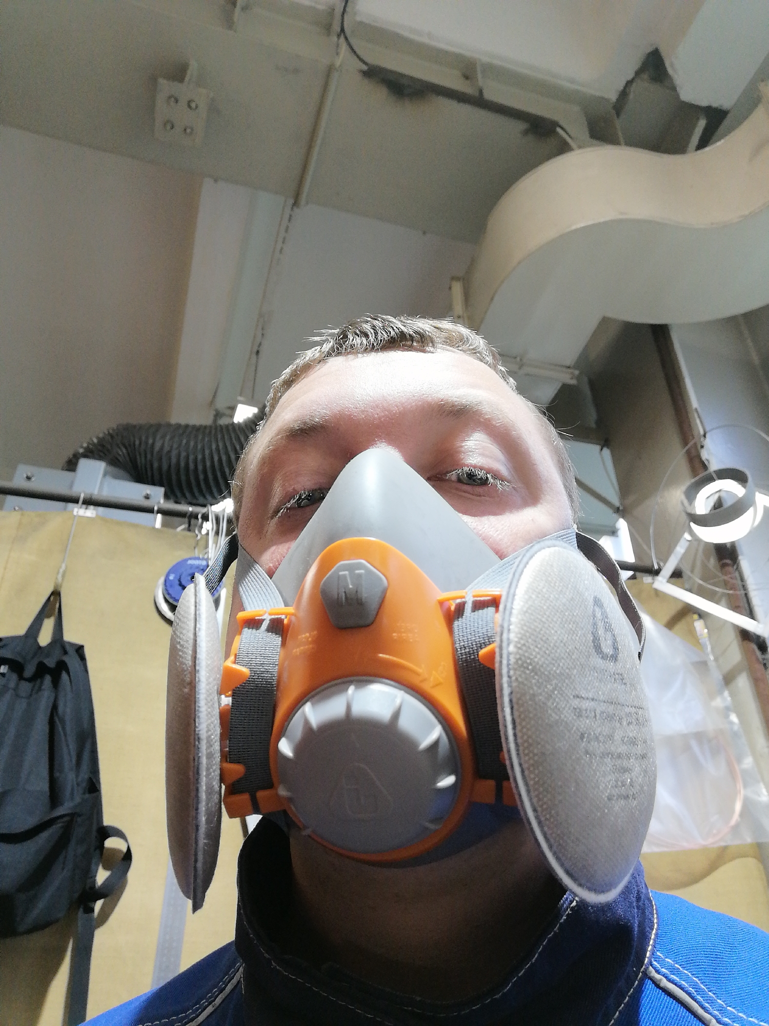 A person wearing a respirator is pictured in a medium shot from a low angle. The person is fair-skinned with short, light-colored hair, with the respirator covering the lower portion of their face. The respirator is gray and orange with gray straps. The person is wearing a blue collared shirt with white stitching. In the background, there is industrial ductwork overhead, as well as a black backpack. There are also several ceiling lights. The location is an industrial or commercial setting, and the city of Unknown, Unknown is unknown.