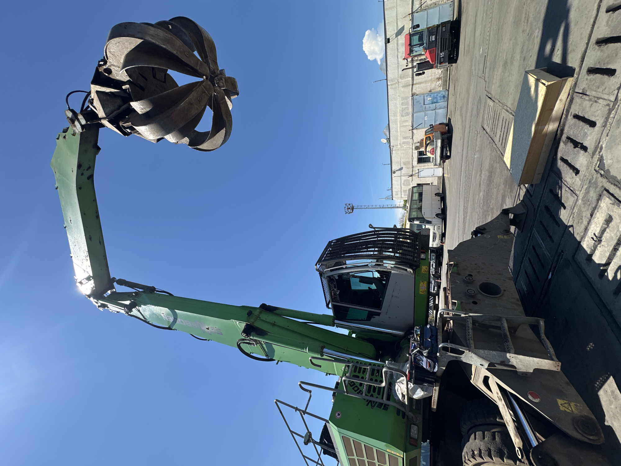 The image shows a large, green industrial grabber machine with a claw attachment against a bright blue sky. The machine is the primary subject, with its extended arm and claw dominating the left side of the frame. The claw appears to be made of metal and has multiple articulated sections. The machine's cab is visible, protected by a metal cage, and the body of the machine shows various hydraulic components and tracks.
In the background, a paved area suggests an industrial or port setting. Several other vehicles are parked or moving in the distance, including a red and black forklift with the number "15" visible, a small white bus, and what appears to be another truck. Buildings with concrete walls and large doors are also present. A single white cloud is visible in the sky. The sun is shining, casting shadows, suggesting it is daytime. The scene appears to be outdoors, possibly in a port or industrial yard. There is no activity taking place in the image; the machine is stationary. Visible text includes "SENNEBOGEN" on the side of the green machine.