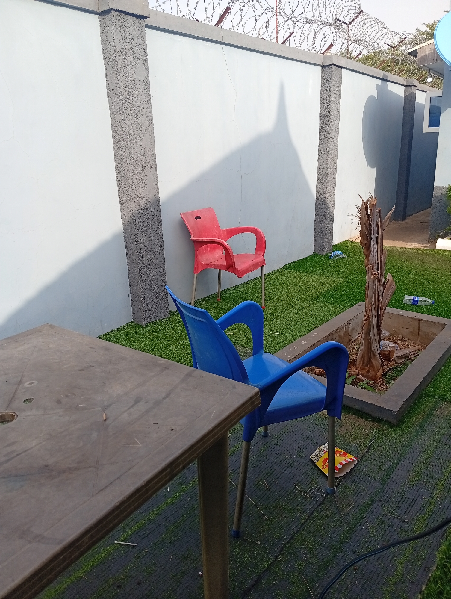 The scene is an outdoor space in Adankolo, Nigeria. A gray plastic table dominates the foreground. Behind the table, a blue plastic chair sits on artificial turf. A red plastic chair is positioned against a white wall with gray vertical support columns. At the top of the wall, there's a razor wire security fence. A planter containing a dead tree is next to the blue chair. Scattered litter, including an empty snack package and plastic water bottles, is on the ground. A shadow is cast on the wall.