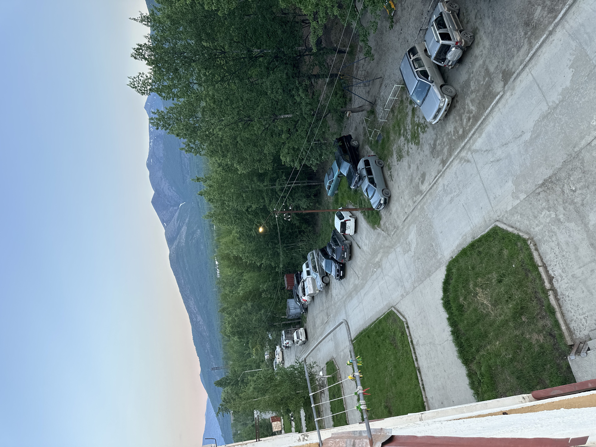 This is a high-angle, full-shot, outdoor, daytime photograph taken in the city of Sinegorye, Russia. The scene depicts a residential area with apartment buildings, a parking lot filled with various cars, and a backdrop of lush green trees and distant mountains. The sky is a pale blue, suggesting either early morning or late evening.
In the foreground, on the left side of the image, a portion of a cream-colored apartment building is visible, with visible signs of wear and tear on its facade, including peeling paint and some discoloration. Attached to the building is a metal structure, possibly a balcony railing, with several colorful, worn-out swing ropes hanging from it. Adjacent to the building is a patch of uneven, grassy lawn.
The main focus of the image is a paved road or driveway that curves through the scene. A row of parked cars lines the right side of this road. The vehicles include several SUVs and sedans, some of which appear to be older models. Notable vehicles include a silver SUV with a spare tire mounted on the back, a dark-colored SUV, and a light gray SUV. Further down the road, more cars are parked, including a white sedan.
Beyond the parking lot and road, a dense forest of tall, green trees dominates the middle ground, stretching towards the horizon. The trees are a mix of deciduous and coniferous species. In the far distance, a range of mountains with snow-capped peaks is visible against the clear blue sky. The lighting suggests it is dusk or dawn, with soft shadows and a diffused light.
The overall impression is of a somewhat rugged, natural environment bordering a developed residential area. The presence of parked cars and apartment buildings indicates a human settlement, while the extensive forest and mountains highlight the natural landscape surrounding the city. There are no visible people actively engaged in any interactions within the frame. The scene is static and quiet, with an emphasis on the built environment set against a natural backdrop.