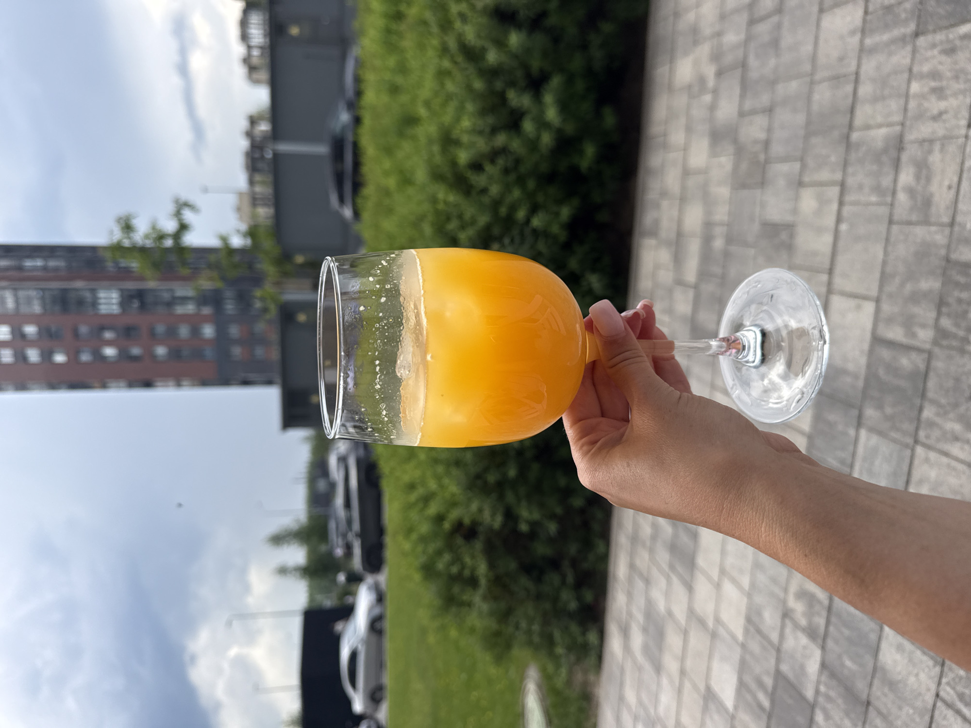 A person's hand is holding a wine glass filled with orange liquid and ice. The glass is on a patio with gray, square tiles. In the background, there is a green hedge, cars, and a tall, reddish-brown building. The sky is cloudy.