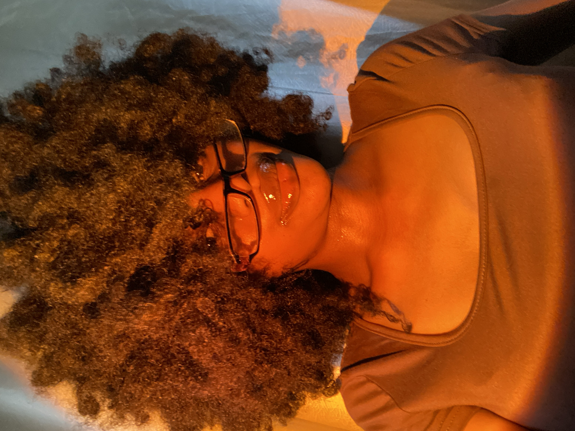 A person with dark skin and large, dark, curly hair, wearing eyeglasses and a light-colored top, is lying on a light-colored surface. The person appears to be sweating. The location is not discernible from the image. There are no visible actions or interactions.
