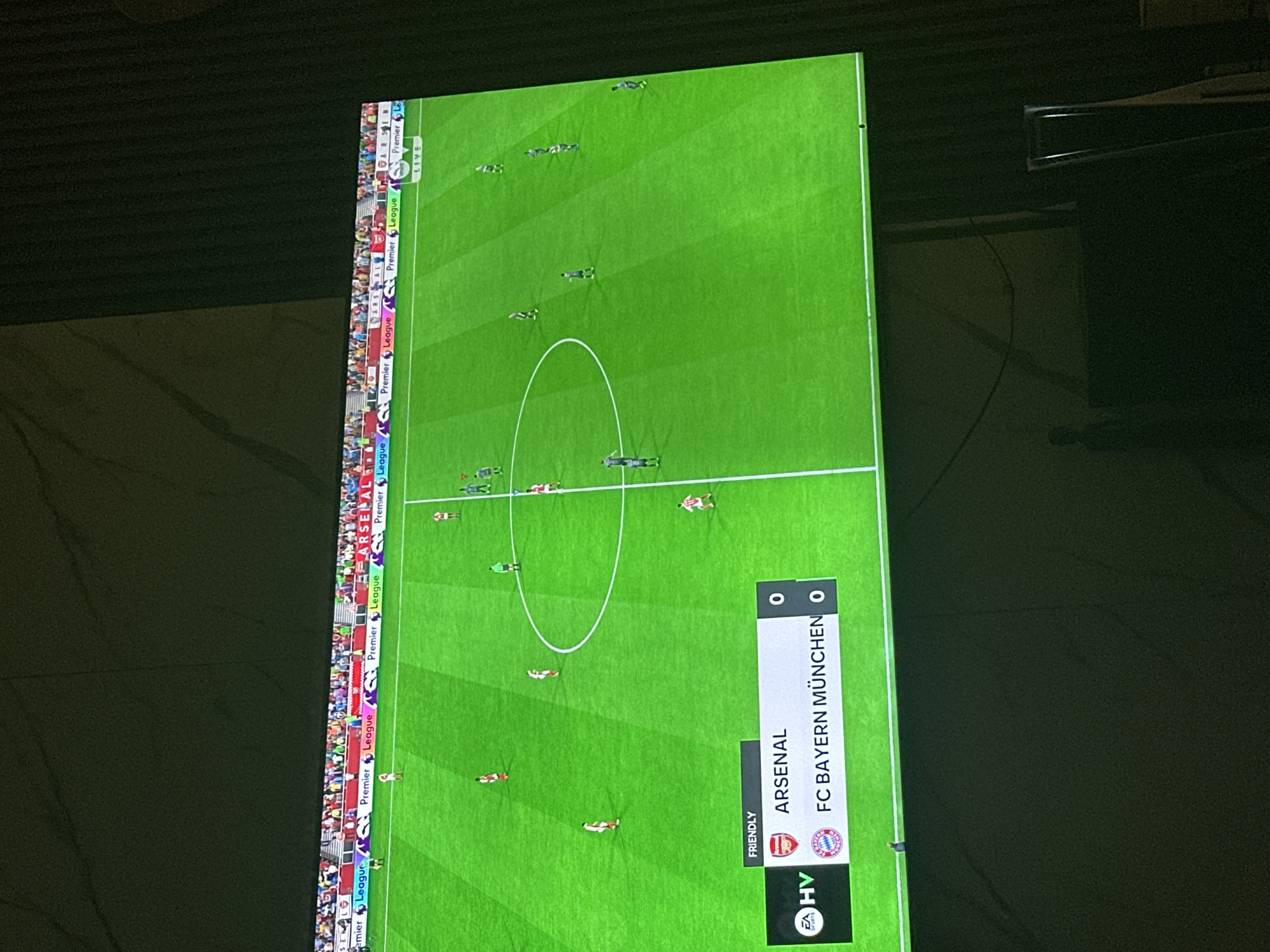 This media file shows a vertical, overhead view of a soccer video game being played on a large screen. The game features two teams, Arsenal and FC Bayern München, with the score currently at 0-0. The setting appears to be an indoor venue, possibly a living room or entertainment space, given the dark background and the presence of what looks like a projector screen. The soccer field is brightly lit, with green turf and white markings. Several digital players are visible on the field, scattered across the pitch. Along the left side of the screen, a banner displays "Premier League" logos, indicating the league the game is simulating. The bottom right corner of the screen shows game information, including the teams' names, scores, and the designation "FRIENDLY". The EA Sports logo is also visible. The overall impression is of a casual gaming session, with no specific time of day or weather conditions discernible. The atmosphere seems relaxed, focused on the on-screen action.