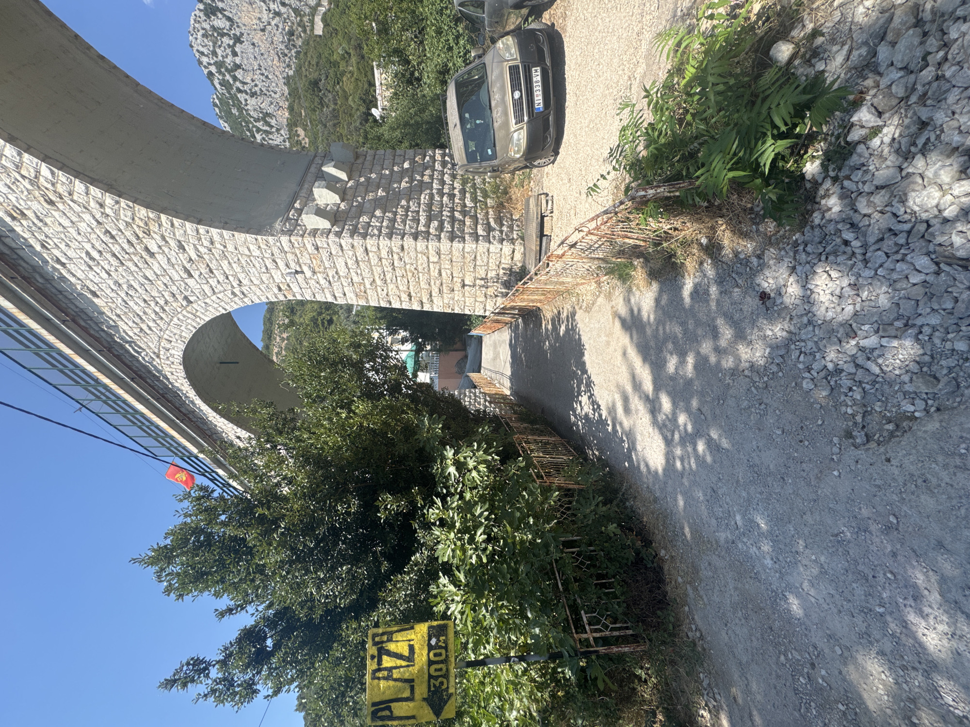 The image depicts a sunny scene in Brca, Montenegro, likely near a beach. A large stone viaduct with multiple arches dominates the upper portion of the frame. A red flag with a golden emblem, possibly the flag of Montenegro, is visible hanging from the viaduct's railing.
In the foreground, a dirt path leads away from the viewer, flanked by lush green vegetation and a rusty, ornate metal fence. To the left, a yellow sign with black lettering reads "PLAZA" and an arrow pointing left, with "300m" below it, indicating the direction and distance to the beach.
A dusty, golden-colored car, a Fiat Doblo with license plate "NI 338-VM", is parked on a gravel area to the right of the path, near the viaduct's support structure. Another car is partially visible behind it. The environment is characterized by a clear blue sky, mountainous terrain in the background, and the presence of trees and shrubs. The overall impression is one of a rural, coastal area on a bright day.