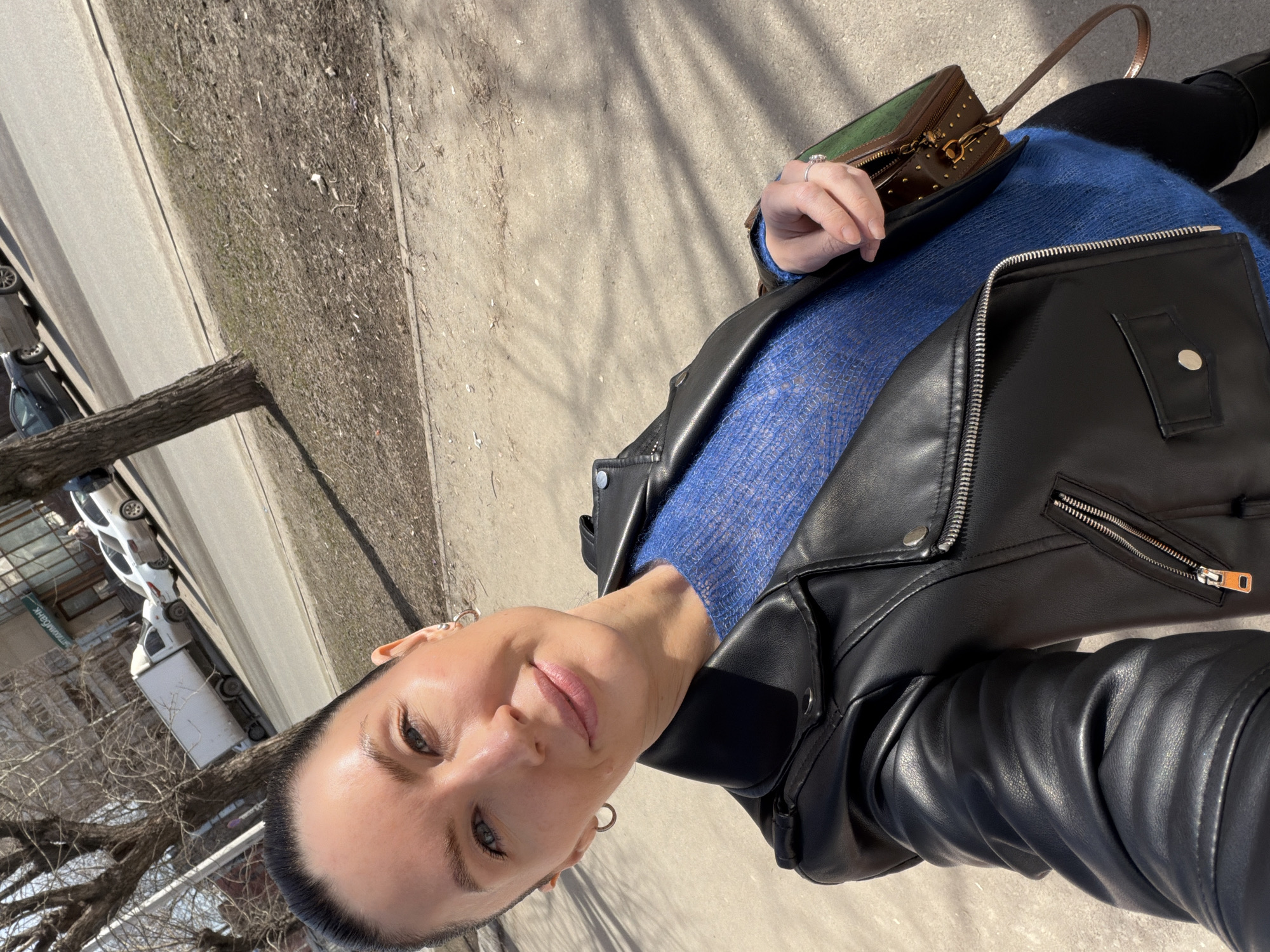 A woman is facing the camera. She has fair skin, short dark hair, blue eyes and is wearing a blue sweater and black leather jacket. She also has a purse in her hand. She appears to be standing on a sidewalk in a city. There is a street visible to the left, along with parked cars. A tree is between the street and sidewalk. Based on the style of architecture, vehicles and foliage visible in the background, it is possible that the location is Perm, Russia.