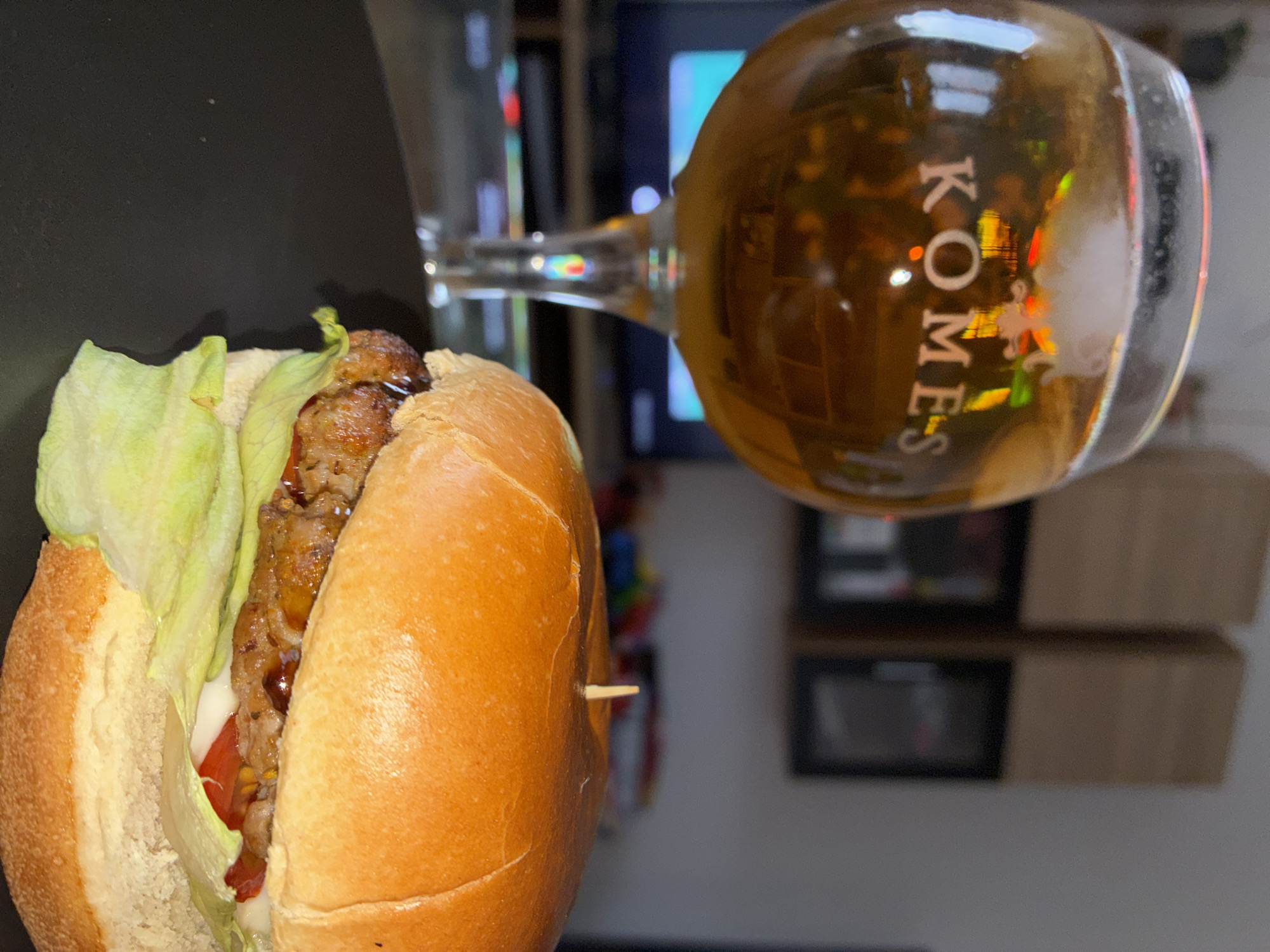 A close-up, angled shot shows a hamburger and a glass of amber liquid. The hamburger is on a dark surface, with the top bun partially covering the patty, lettuce, and a slice of tomato. A toothpick protrudes from the side of the bun. To the right, a stemmed glass filled with a golden beverage rests on its side. The glass displays text that reads "KOMES". The background is blurred, showing what appears to be a television screen with colorful reflections and some shelving units. The lighting suggests an indoor setting, possibly at night or in a dimly lit room. The overall scene conveys a casual, relaxed atmosphere, perhaps a meal enjoyed while watching television. The visible text indicates the beverage is likely a beer or similar drink. Given the location context of Toruń, Poland, "Komes" could refer to a specific brand of beer popular in the region.