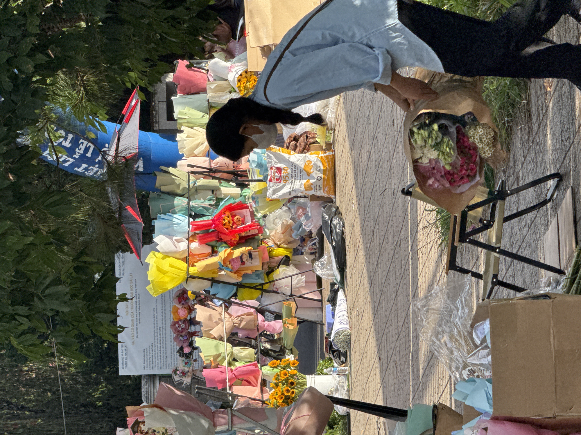 The image depicts an outdoor scene likely in Lâm Viên - Đà Lạt, Vietnam, characterized by a street-level view of a flower stall. In the foreground, a person wearing a light blue shirt and dark pants, with a white face mask, is holding a large bouquet wrapped in brown paper. The bouquet contains a variety of flowers, including pink and white blooms. To the left of this person, a portable chair with a metallic frame and brown fabric is visible. Behind the person, a rack displays numerous colorful bouquets wrapped in paper in various shades of pink, yellow, green, and blue. A white sign with Vietnamese text is mounted on a wall or structure in the background. Several bags of what appear to be pet food are also present near the flower stall. The ground is paved with hexagonal tiles, and greenery can be seen on the right side of the image. The overall atmosphere suggests a commercial street scene with a focus on the sale of flowers.
