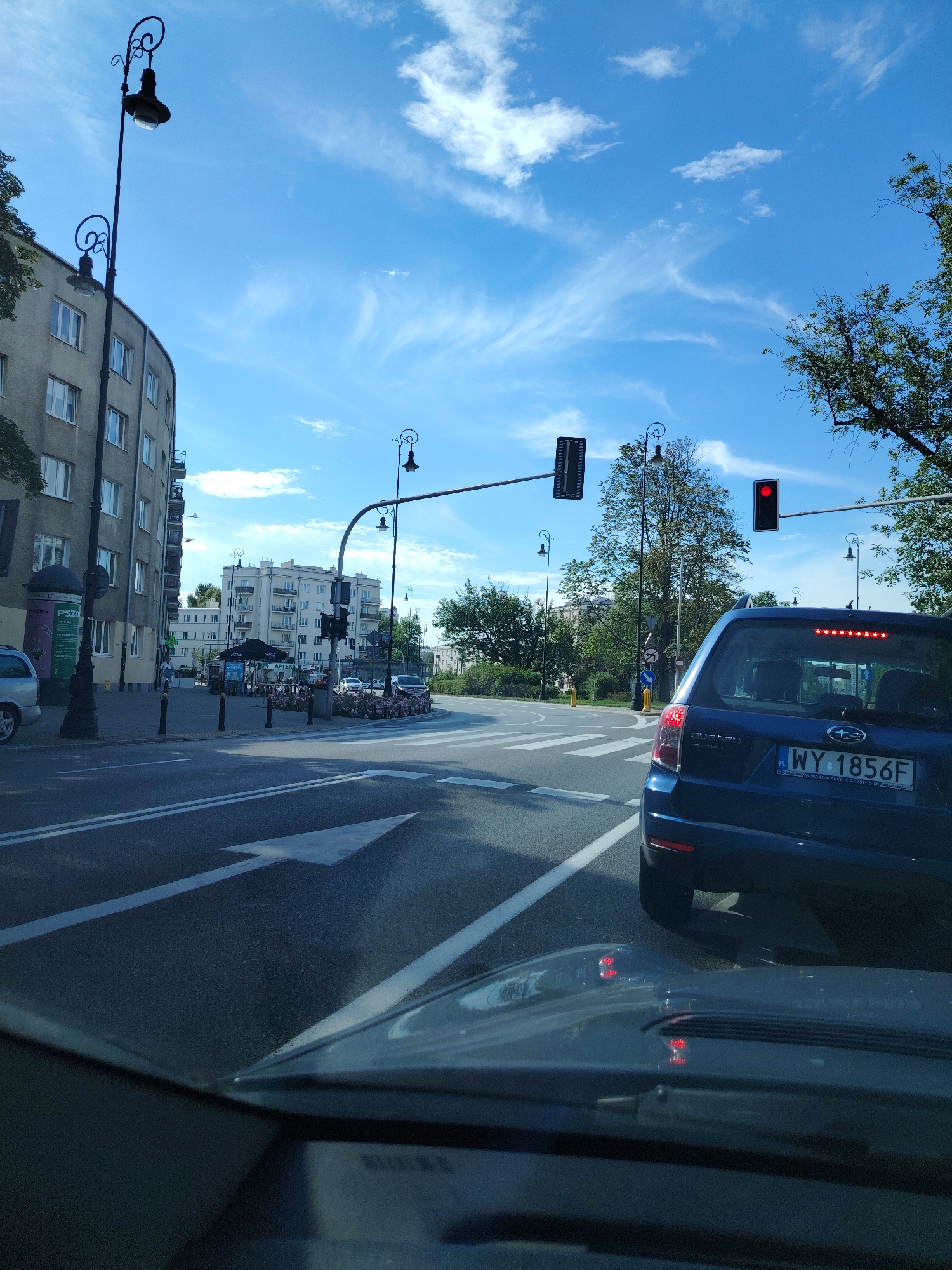 This is a street-level, eye-level, vertical, full shot of a city intersection in Warsaw, Poland, taken from the perspective of a driver. The sky is bright blue with wispy white clouds. A traffic light with a red signal is visible, indicating a stop. In the foreground, the hood of the car the photo is being taken from is visible. Behind it, a blue Subaru Forester with the license plate WY 1856F is stopped, facing away from the camera, also at the intersection. The car's brake lights are illuminated. The road has white lane markings, including a directional arrow pointing forward and to the left. On the left side of the road, a multi-story building with a grey facade and numerous windows lines the street. A lamppost with an ornate design stands on the sidewalk. Further down the street, there are more buildings, some with awnings, and trees. The scene suggests a typical urban environment during the daytime.