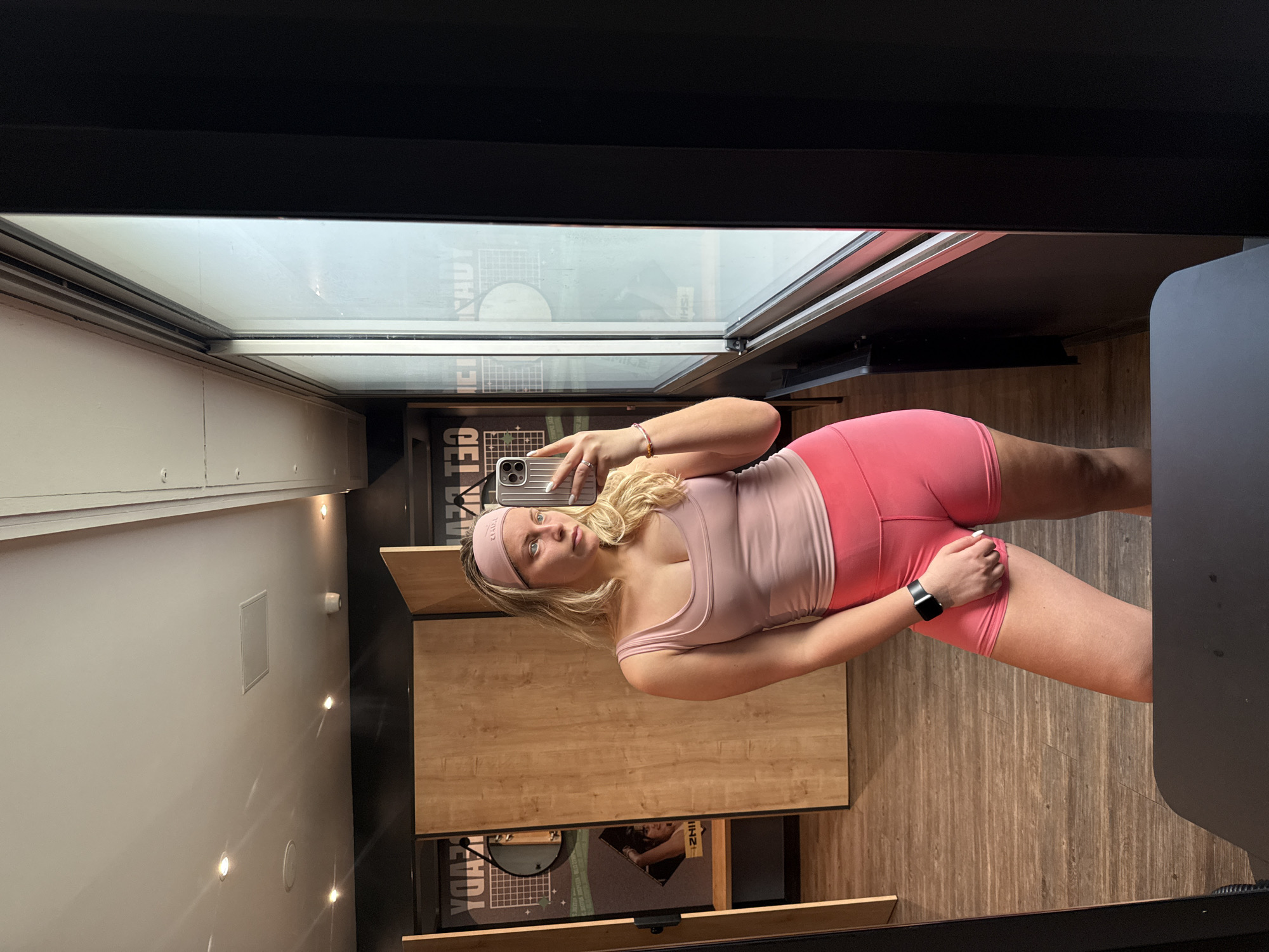 A young woman wearing a pink headband, a light pink tank top, and bright pink shorts is taking a selfie in front of a mirror. The mirror is framed in black and reflects the interior of a space that appears to be a locker room or dressing area with wood-paneled walls and a window with natural light coming through. There are graphics and text visible on the wall behind her, possibly related to branding or advertisements. The woman is holding a phone with a metallic case to take the picture and a black smartwatch is visible on her wrist. The location may be in a gym or fitness studio in Paris, France, given the dressing room context, but there's not enough location data in the image to confirm.