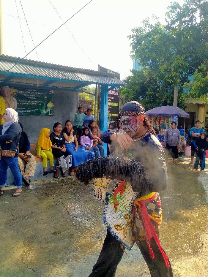 An outdoor scene in Sepat, Indonesia, depicts a performance with an audience. In the foreground, a male performer with elaborate white and black face paint, a black headscarf, and dark clothing holds a decorated, horse-head-shaped prop with a black mane and colorful fabric sashes. Vapor or smoke emanates from his upper body. The ground is concrete, appearing wet in areas. To the left, a group of approximately 10-12 individuals, primarily women and children, are seated on a low concrete bench under a corrugated metal roof, observing the performance. A banner on the adjacent wall displays Indonesian text, including "KARANGTARUNA MIRABUMI" and "DESA RENDINGAN." To the right, several more individuals stand near a purple umbrella-covered stall. The background features green trees and a light blue building. The scene is illuminated by bright daylight.