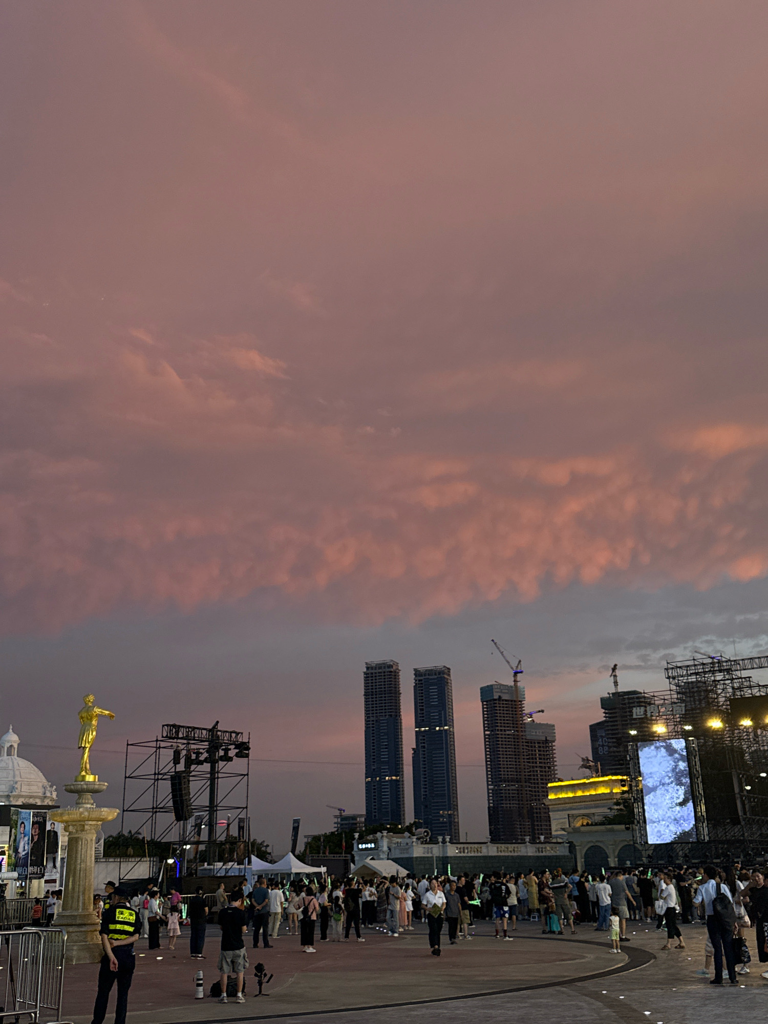 The image depicts an outdoor gathering of people in what appears to be a public square at dusk. The sky is filled with dramatic pink and purple clouds, suggesting a sunset. In the foreground, a golden statue stands on a pedestal, with a security guard in uniform standing to its left. A crowd of people are milling about in the middle ground, some holding glowing green sticks, indicating a possible event or concert. Behind the crowd, there is a stage structure with speakers and lighting equipment. In the background, several modern skyscrapers pierce the sky, with one featuring a large LED screen displaying an image. Construction cranes are also visible, suggesting ongoing development in the area. The scene appears to be taking place in a bustling urban environment, likely within Nanshan District, China, given the architectural style of the buildings.