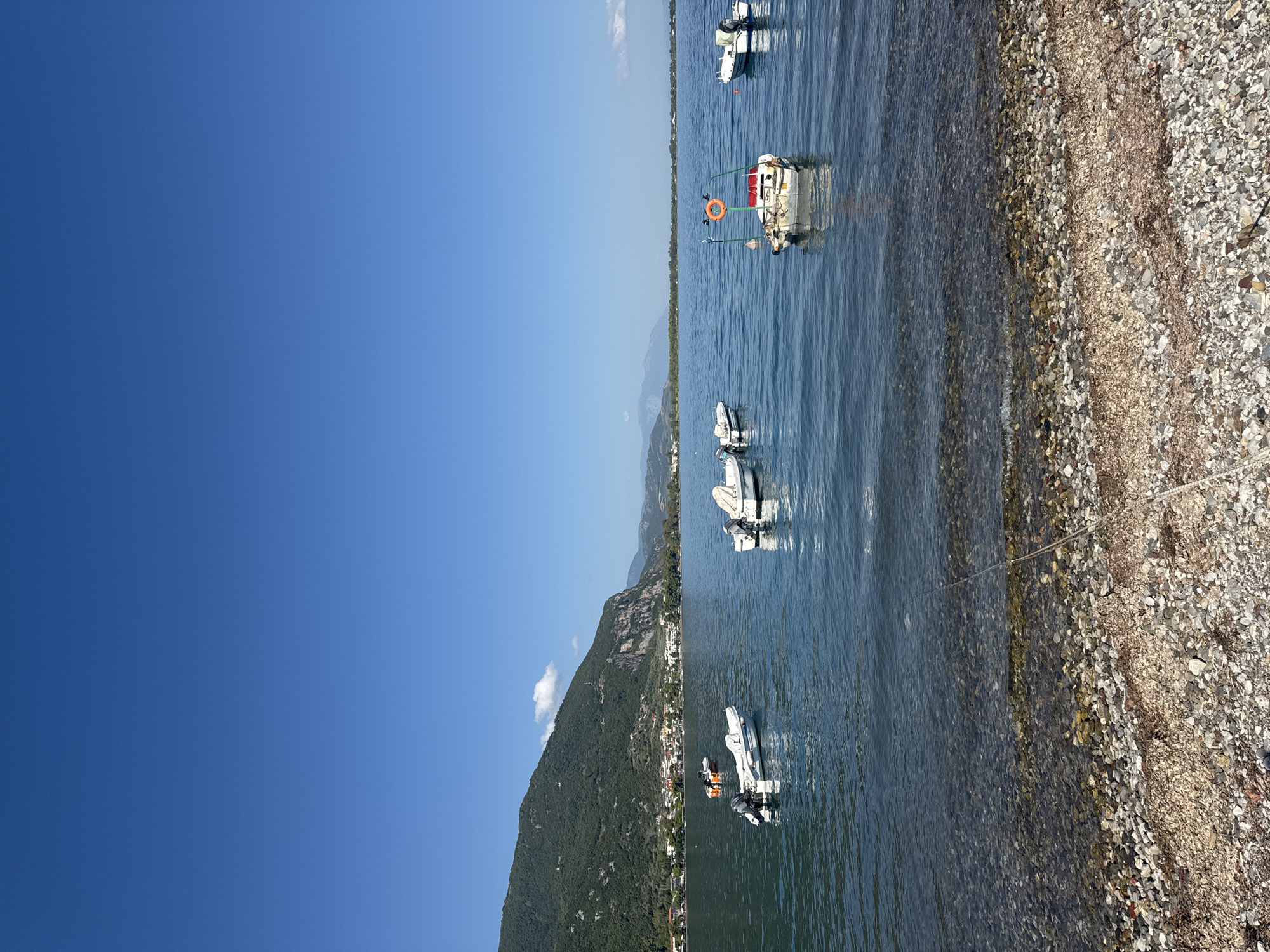 The image depicts a serene coastal scene in the Municipal Unit of Kamena Vourla, Greece. The foreground is a pebbled beach with scattered seaweed and a visible rope extending into the water. The water is clear and calm, revealing the submerged pebbles. Several small motorboats are anchored in the bay, some closer to the shore and others further out.
In the mid-ground, a line of buildings forms a coastal settlement, backed by a densely vegetated, steep mountain that dominates the left side of the frame. The mountain's slopes are covered in green foliage, with rocky outcrops visible in places. The sky is a clear, vibrant blue, with a single, small white cloud near the mountain peak.
Further in the distance, a hazy mountain range stretches across the horizon, meeting the sea. The water is a deep blue, reflecting the clear sky. The overall impression is one of tranquility and natural beauty. There are no people visible in the image.