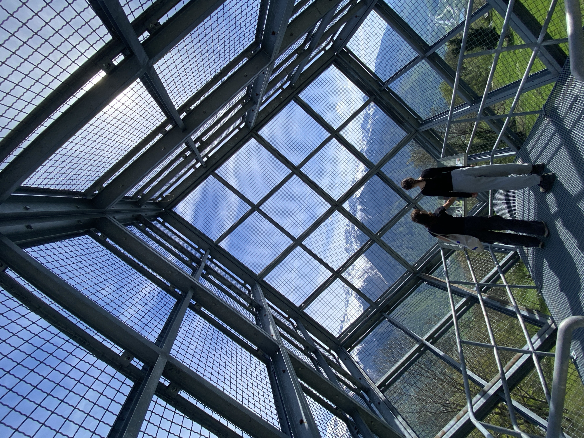 This low-angle photograph captures the interior of a modern observation structure, likely a skywalk or viewing platform, situated in Saint-Georges-d'Hurtières, France. The scene is dominated by a robust framework of dark grey metal beams forming a grid pattern, supporting both transparent panels and metal mesh.
The camera is angled upwards and outwards, revealing a bright blue sky with scattered white clouds through the structure's ceiling panels. The sun shines intensely, creating a noticeable lens flare in the upper left quadrant. The floor of the structure consists of a metal grid and transparent panels, offering a direct view to the landscape below.
Two individuals are present on the transparent floor on the right side of the image. One person, dressed in a black long-sleeved top, light grey trousers, and dark shoes, stands with their back largely to the viewer, seemingly observing the distant vista. A white carrier bag, possibly with the words "BETTER SHAPE" visible, is held near them. A second person in dark attire is partially visible beside the first.
Through the transparent floor and side panels, a verdant, mountainous landscape unfolds far beneath the structure, featuring green grassy slopes and trees. The photograph conveys a clear, sunny day, highlighting the structure's impressive height and the expansive natural beauty of its surroundings.