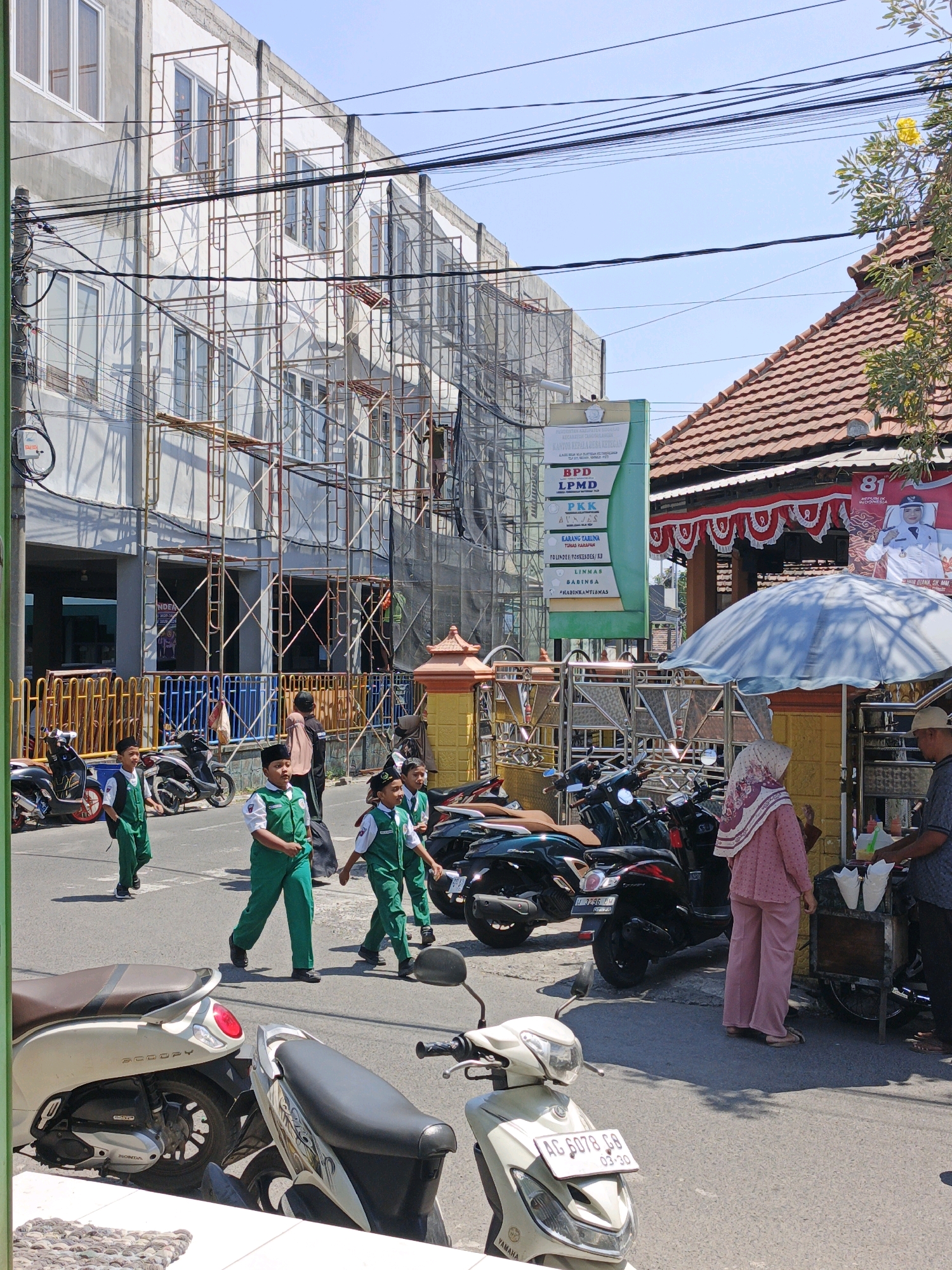 The image depicts a street scene in Ketegan, Indonesia, on a bright, sunny day. In the foreground, several motorcycles are parked. To the left, a cream-colored Honda Scoopy is prominently visible. Further back, other motorcycles, including a dark-colored one with a red taillight, are parked.
The middle ground features a group of boys in green school uniforms walking on the sidewalk and street. They are wearing white shirts and black hats or caps. Behind them, a woman in a pink hijab and matching outfit stands near a food cart, and a man in a dark shirt and cap appears to be interacting with her. The food cart has an umbrella providing shade.
The background shows a multi-story building under construction, with scaffolding covering a significant portion of its facade. The building has numerous windows. A tall green billboard with text in Indonesian is positioned near the building. To the right of the building, a traditional Indonesian-style roof with red tiles is visible. Overhead, a tangle of electrical wires crisscrosses the sky. The overall environment suggests an active urban area with ongoing development and daily life unfolding.