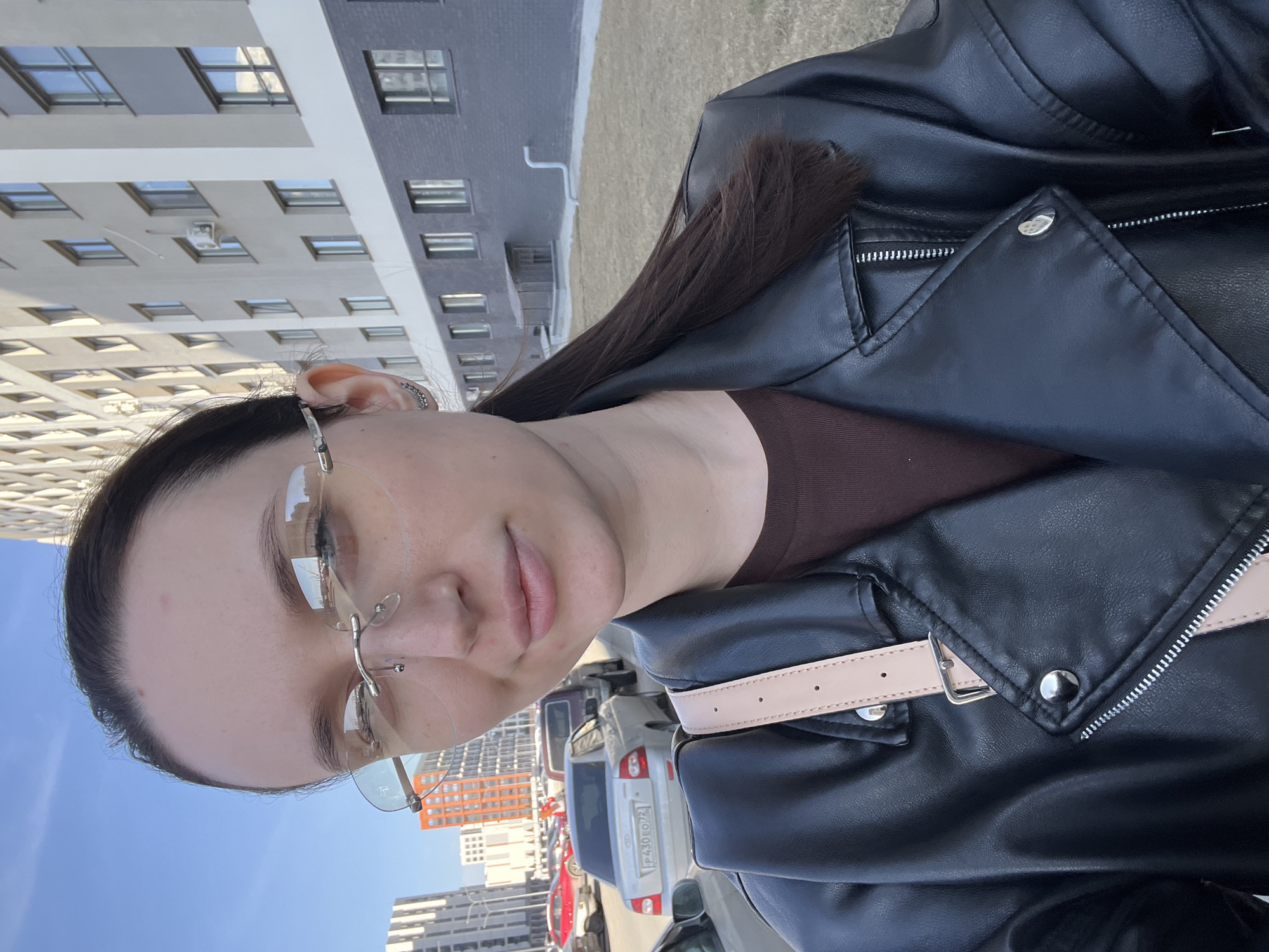A person with dark hair and eyeglasses is facing the camera, wearing a black leather jacket and a brown top. The background shows buildings and cars, indicating an urban environment. The license plate of a silver car is partially visible. Based on the license plate code "430", the location can be identified as Tyumen, Russia.