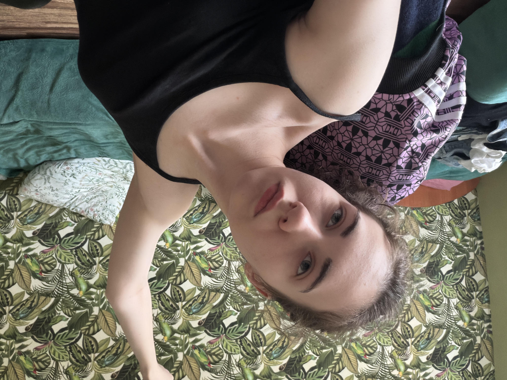 The image shows a young woman with light skin and brown hair, wearing a black tank top. She is indoors, with a wall covered in a tropical-themed wallpaper featuring green leaves and parrots. To her left, there is a bed with a teal-colored blanket and a pillow with a floral pattern. There are also some clothes piled on a chair or surface next to her. The woman's right arm is raised and bent at the elbow. The lighting suggests indoor ambient light. There is no indication of location within Zavodoukovsk, Russia.