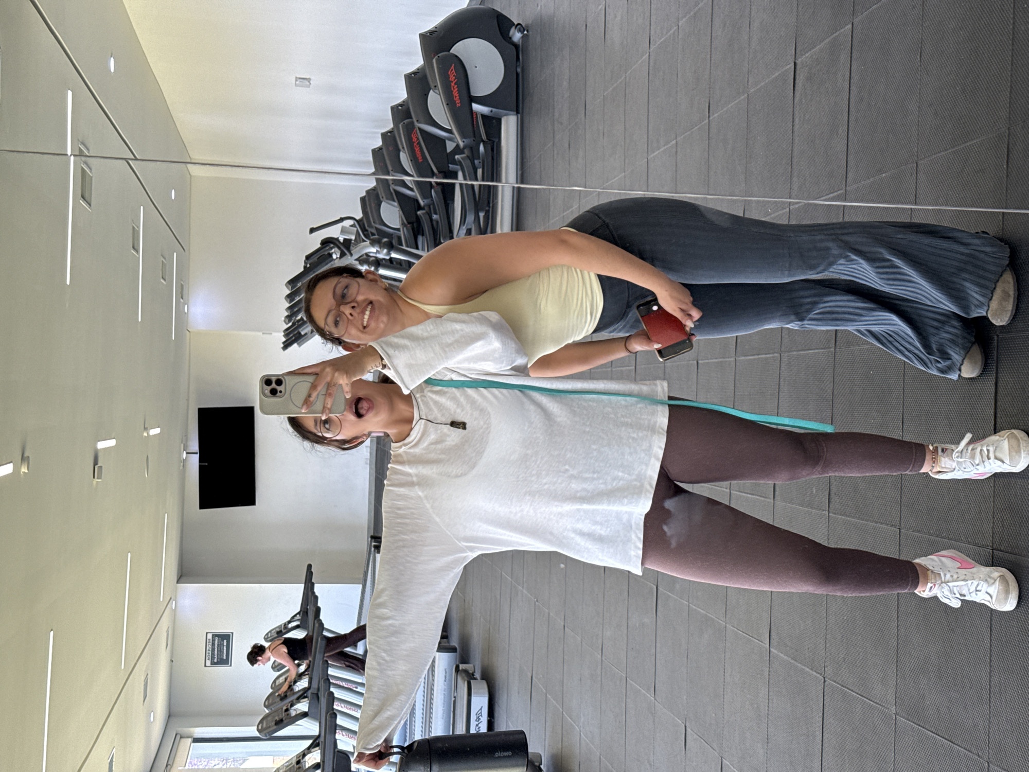 Two women are posing in front of a gym mirror, reflected in the mirror. One woman is wearing a yellow tank top and jeans, holding a phone. The other woman is wearing a white long-sleeved shirt and brown leggings, also holding a phone for a selfie. Behind them are rows of exercise equipment and weights. The location is in San Andrés Cholula, Mexico.