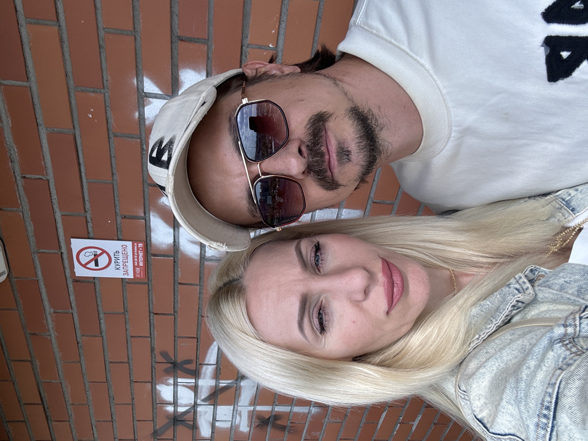 A selfie shows a man and a woman posing against a brick wall. The woman, on the left, has long blonde hair and is wearing a denim jacket. The man, on the right, is wearing a baseball cap and sunglasses. A sign on the wall reads "КУРИТЬ ЗАПРЕЩЕНО" (Smoking is prohibited) and includes a no-smoking symbol. Graffiti is visible on the wall below the sign. The background appears to be an outdoor urban setting.