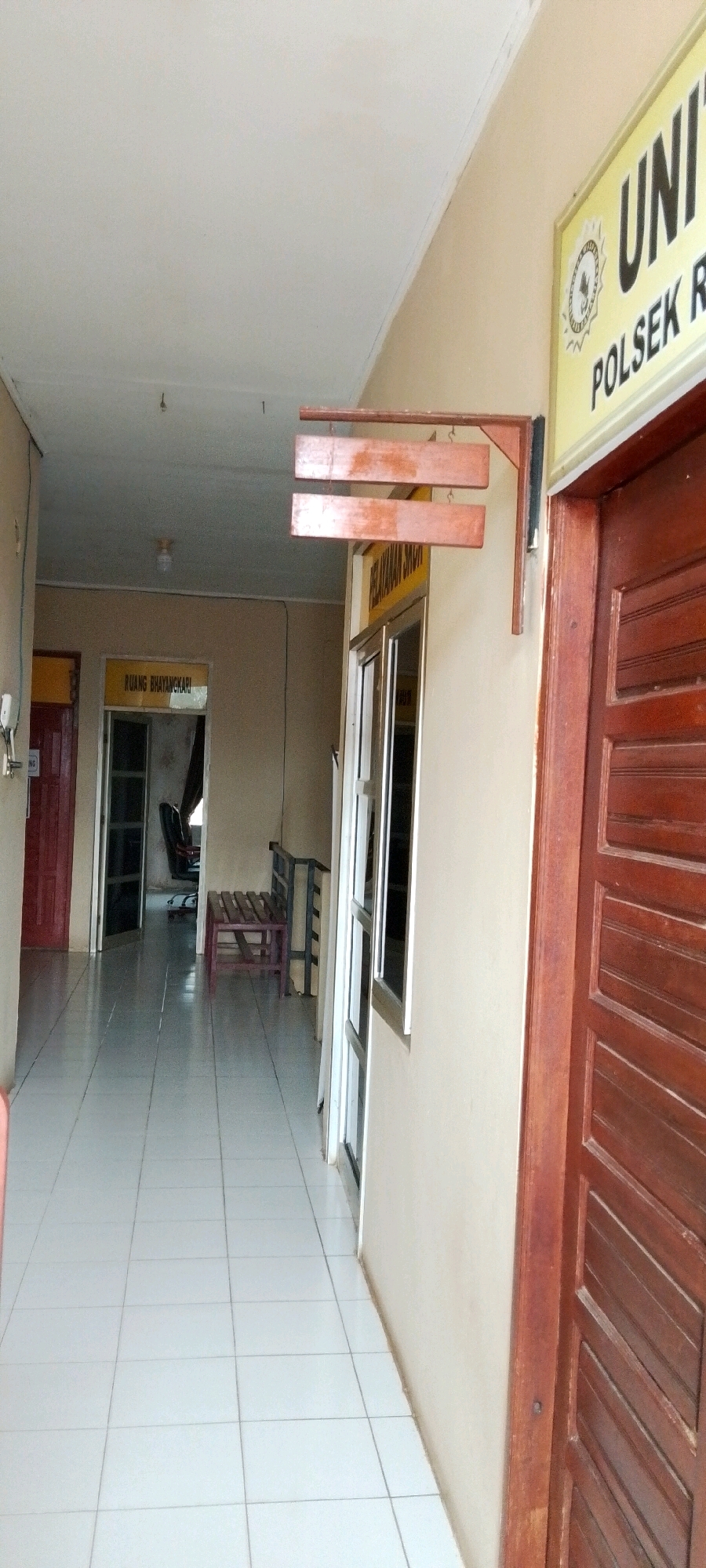 The image shows an interior view of a hallway in a building in Unknown, Indonesia. The hallway has white tiled floors and beige walls. On the left side of the hallway, there are two red doors and a doorway leading into a room with a chair and desk. Further down the hallway, there is a wooden bench. On the right side of the hallway, there is a wooden door and a window with a yellow sign above it that reads "UNIT POLSEK R". There is also a wooden structure with three planks hanging from it mounted on the wall above the window. The lighting in the hallway appears to be artificial, with a light fixture visible on the ceiling. The overall scene suggests a public building, possibly a police station or government office.