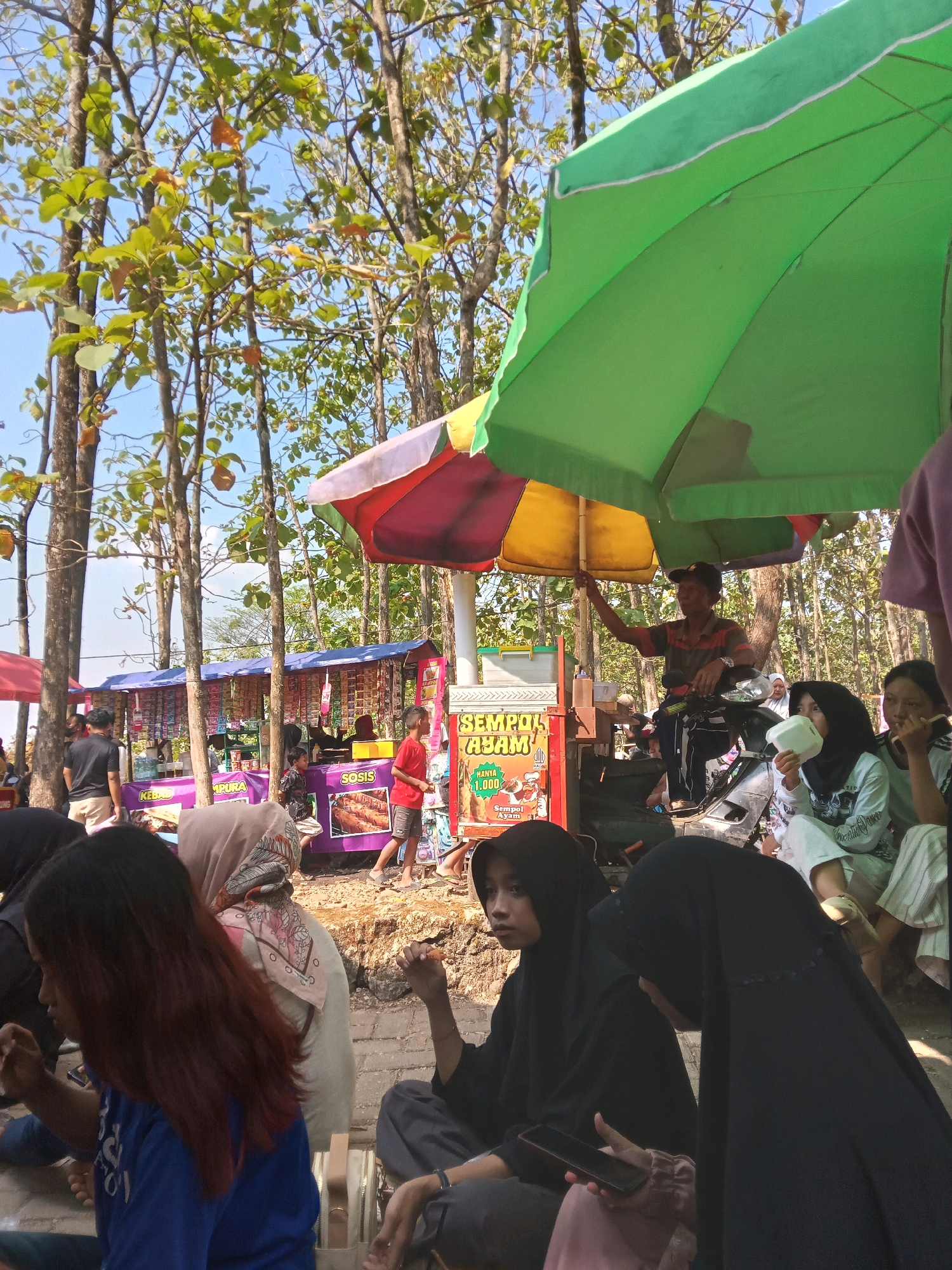 This image captures a vibrant outdoor market scene, likely in Kedunggudel, Indonesia, under a canopy of trees. A food stall, marked with "SEMPOI AYAM," is prominent, with a vendor attending to customers. Several people are gathered around, some sitting and eating, while others stand and observe. The market appears to be a bustling hub of activity, with various goods displayed on stalls in the background. The environment is rustic, with dirt pathways and the natural presence of trees indicating an outdoor setting. The atmosphere suggests a casual, community-oriented gathering.