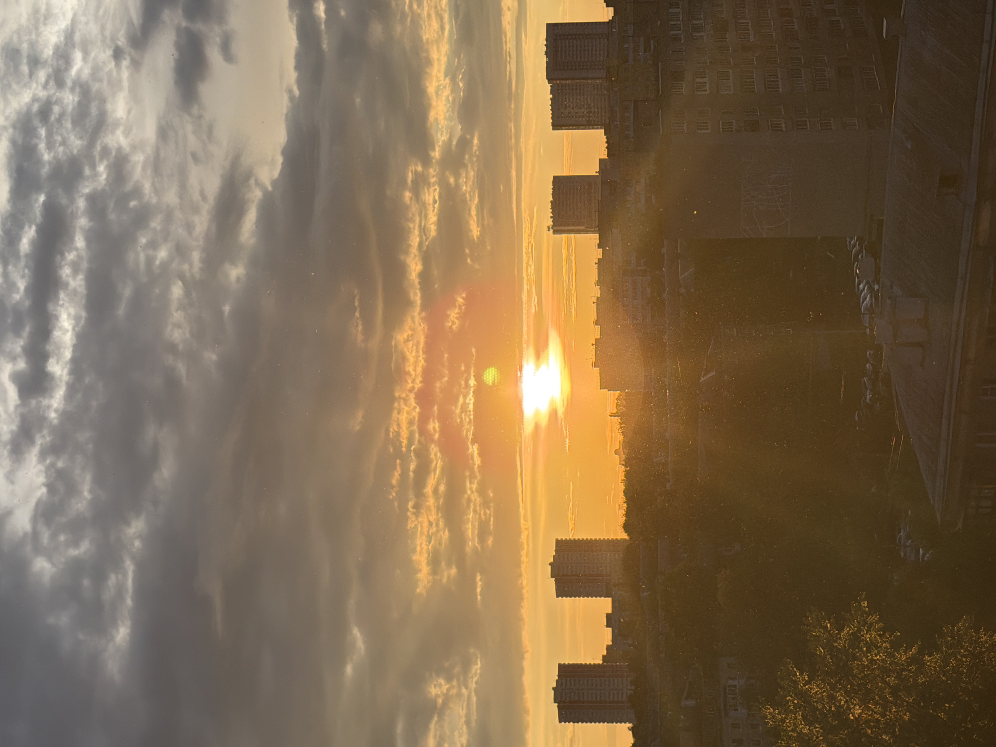 The image displays a dramatic sunset or sunrise over an urban landscape, viewed as if looking out from a window. The photograph is oriented vertically, showing the sky at the top and the city below, with the sun situated intensely bright on the right side near the horizon.
The sky occupies the upper portion of the frame, transitioning from dense, dark gray clouds on the left to streaky, golden, and orange clouds brightly illuminated by the sun on the right. The sun itself is a powerful golden orb, producing a significant lens flare and radiant light rays that stretch across the sky and atmosphere.
Below the illuminated sky, the silhouette of a city skyline is visible. Several tall, rectangular buildings, likely apartment blocks or offices, stand starkly against the bright backdrop. Details of their structures are largely obscured by the backlighting, though some window patterns can be faintly discerned. Further down, in the lower right, dark foliage from trees is evident, catching some of the warm light.
A notable feature throughout the image is the presence of numerous tiny, bright specks, resembling dust, pollen, or perhaps illuminated water droplets, suspended in the air. These particles are particularly visible in the sun's rays and against darker areas, contributing to a hazy or atmospheric quality.
The overall impression is one of a serene yet powerful natural event unfolding over a metropolitan area. The time of day is clearly either sunrise or sunset, characterized by the low position of the sun and the warm, golden hues dominating the scene. There is no visible text in the image.