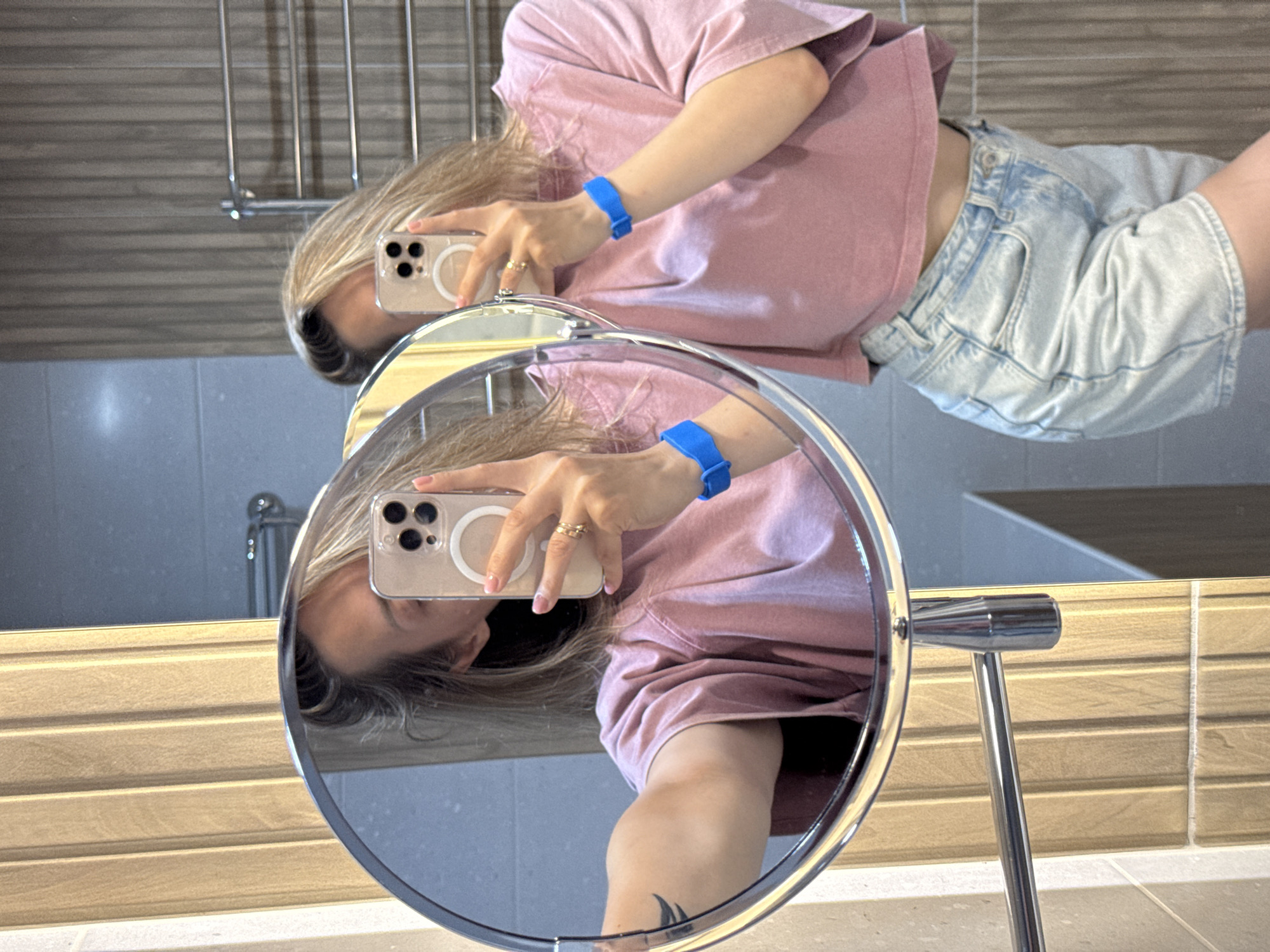 A woman is taking a mirror selfie in a bathroom in Nebug, Russia. She is wearing a pink t-shirt and light blue denim shorts. She has blonde hair and is wearing blue wristbands on both wrists and rings on her fingers. The bathroom has wooden-paneled walls and tiled surfaces. A towel rack is visible in the background.