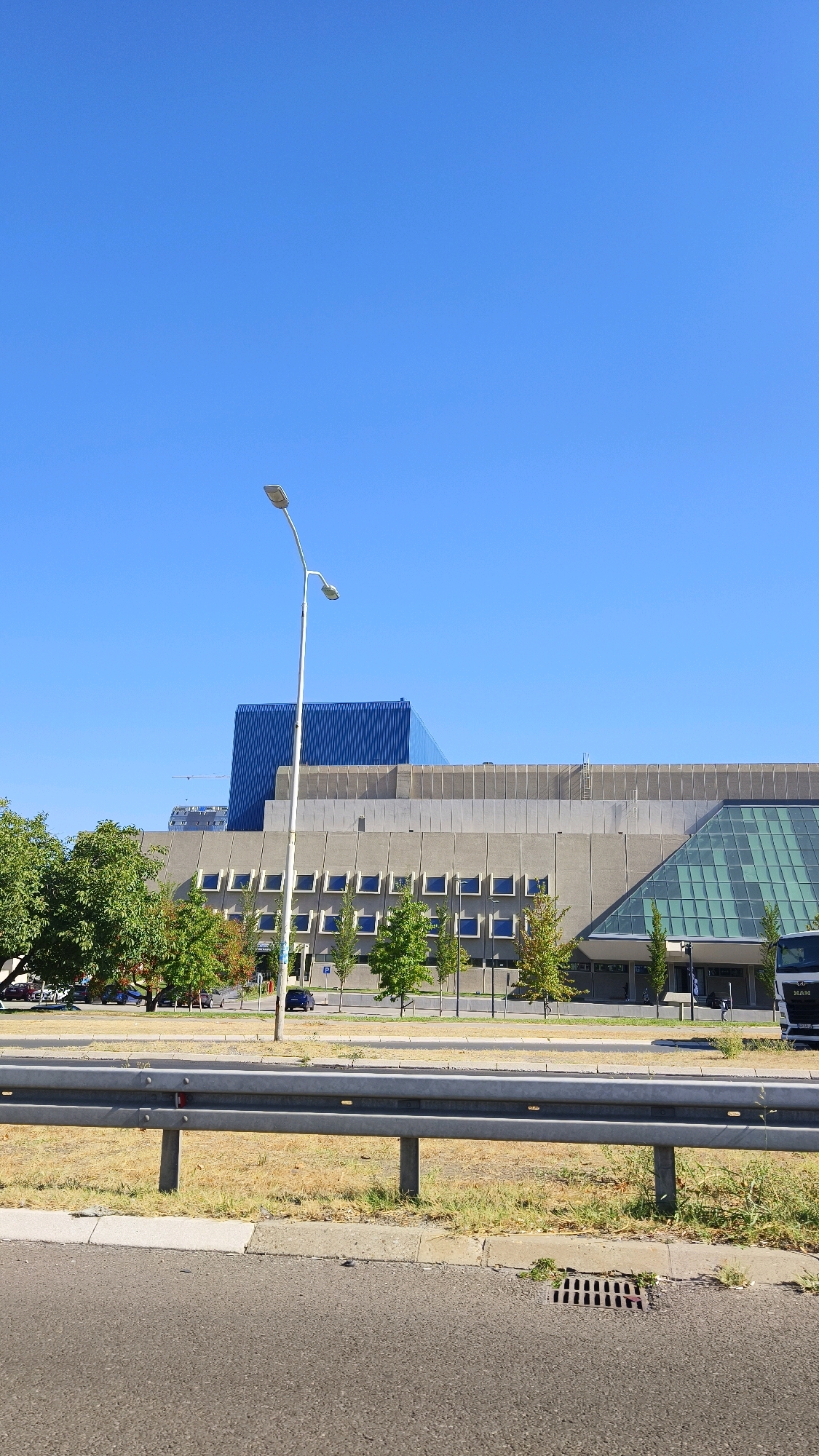 The image captures a daytime outdoor scene in Belgrade, Serbia, featuring a modern building complex and a street. Dominating the sky is a clear, bright blue sky. In the foreground, a grey metal guardrail separates the asphalt road from a grassy area with dry patches. A street light with two lamps stands tall on the left side of the frame.
Behind the guardrail, a long, multi-story building with a concrete facade and rows of square windows is visible. A distinctive blue, angular structure is situated behind and to the left of this main building. To the right of the concrete building, a section with a large, sloped glass roof is apparent. Trees with green foliage line the area in front of the buildings.
Several vehicles are present on the road and in the vicinity of the buildings, including a dark blue car, a silver car, and a white truck on the far right. A street sign with a "P" indicating parking is also visible. The overall impression is one of an urban environment, possibly an administrative or business district.