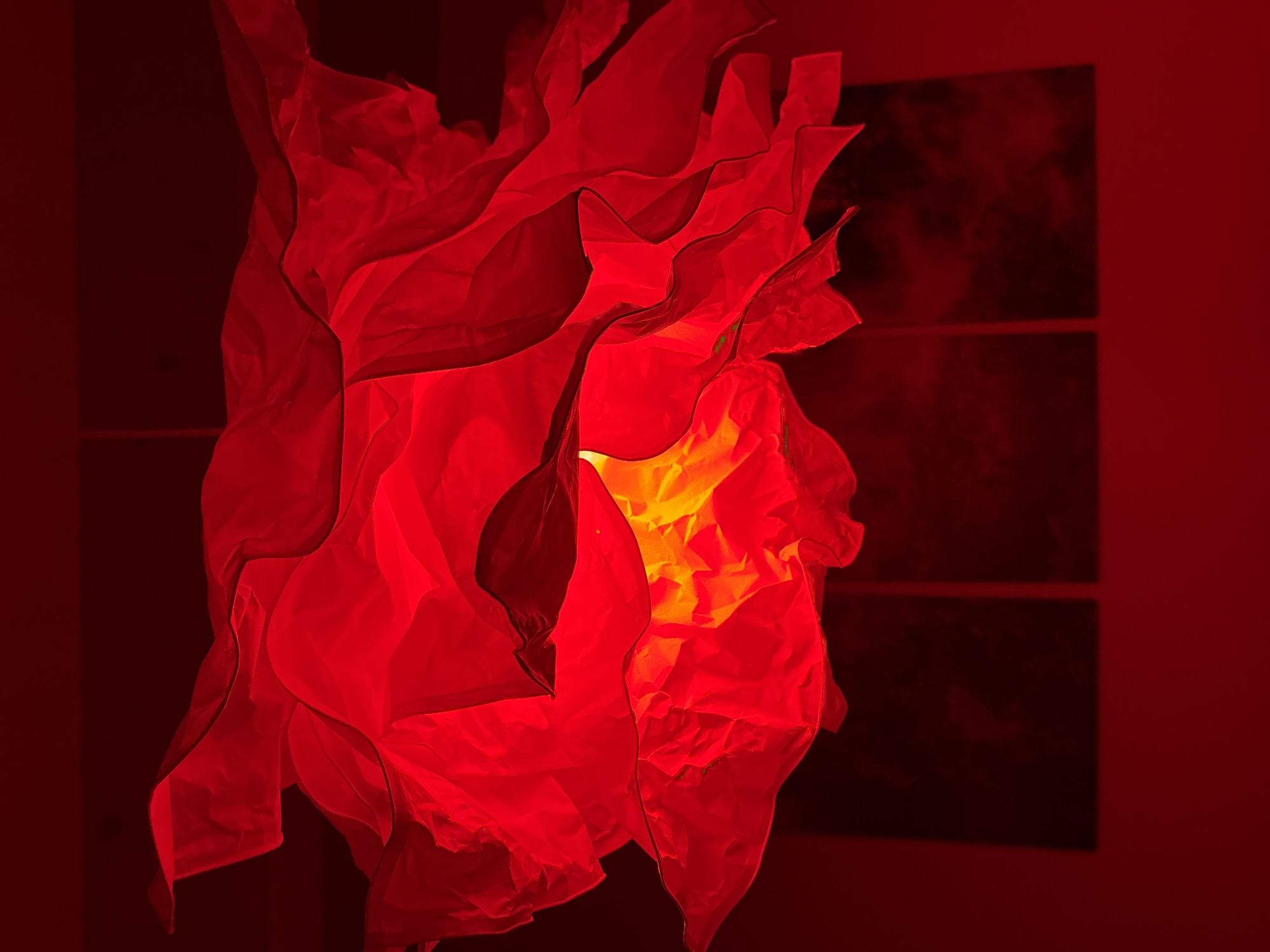 The image depicts an interior scene in Chaikovsky, Russia, dominated by shades of red. A large, abstract light fixture hangs centrally from a dark ceiling, crafted from multiple undulating and ruffled layers of translucent red fabric or paper. An internal light source emits a warm yellow-orange glow, illuminating the center of the fixture from within and casting reddish light outwards. Below the fixture, a deep crimson wall is visible, on which three dark, rectangular panels are mounted horizontally. These panels appear as abstract decorative elements or artworks, exhibiting subtle patterns or textures in darker tones against the wall. No individuals, actions, or interactions are present in the scene.