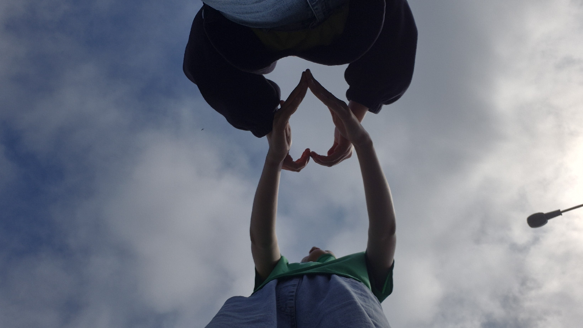 The image, captured in Chaikovsky, Russia, presents a low-angle view looking up towards a cloudy, bright sky during the daytime. Two individuals are featured, with their bodies partially visible from a worm's-eye perspective.
At the top of the frame, the lower half of one person is shown, clad in blue jeans and a dark-colored garment resembling a hoodie or jacket. Below them, another person is seen from the waist up, wearing a green t-shirt and light-colored denim shorts or a skirt. Their arms are extended upwards, and their hands meet to form a clear heart shape against the sky, symbolizing love, affection, or connection.
The sky is a mix of light blue and soft white clouds, with a noticeably brighter area towards the right, suggesting the general direction of the sun. A dark, silhouetted street light fixture is partially visible on the far right edge of the frame, hinting at an urban or suburban outdoor setting. No text is visible in the image.
