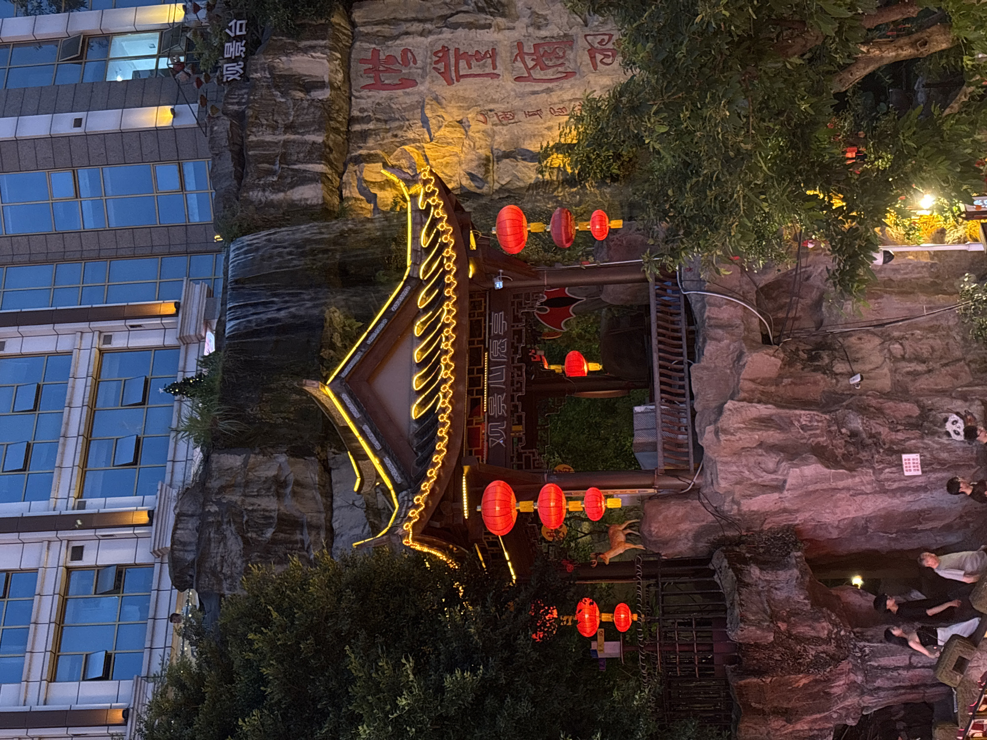 In the Yuzhong District of China, a scene unfolds featuring a traditional Chinese pavilion illuminated by strings of golden lights and red lanterns. The pavilion is situated amidst a rocky landscape with a man-made waterfall cascading down a stone cliff face. A modern building with blue-tinted windows stands in the background, contrasting with the traditional architecture. A small deer figurine stands atop a rock near the pavilion. Below, a group of people are visible, some walking down a path. Chinese characters are inscribed on the rock face. The image appears to be taken at dusk or night, with artificial lighting playing a significant role in the scene.