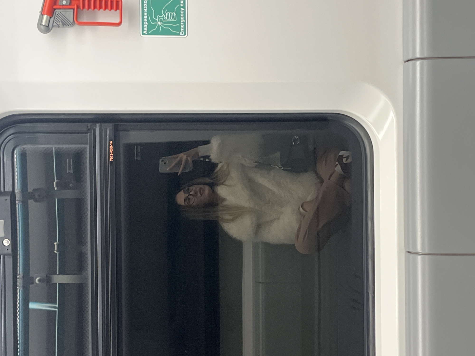 This image captures a reflection of a young woman taking a selfie inside what appears to be a modern subway or train car in Sofia, Bulgaria. The image is rotated 90 degrees clockwise, presenting the scene horizontally.
The woman, with long light-colored hair and wearing glasses, is seen reflected in the train window. She is dressed in a fluffy, light-colored top and light brown pants, holding a light-colored smartphone in her right hand to take a self-portrait. Her posture suggests she is either seated or crouched inside the carriage.
The interior of the train is clean and brightly lit, featuring white and light gray panels. Visible above the window are a red emergency handle and a green "Emergency exit" sign. The sign clearly displays text in both Cyrillic ("Авариен изход") and English, indicating the location. A small identifying sticker with the code "T013-R3B-14" is also present on the window frame.
The dark reflection outside the window suggests the train is either in a tunnel, at a station with a dark background, or traveling during nighttime. The overall impression is one of a passenger engaging in a common activity while commuting on public transport.