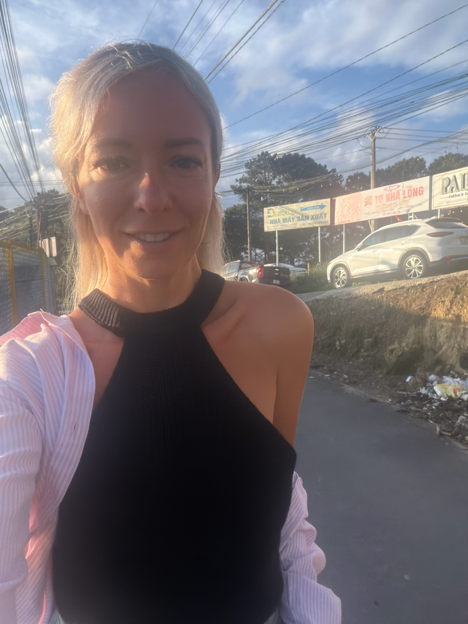 A woman with blonde hair, wearing a black top and a pink and white striped shirt, smiles at the camera while standing on a road. Behind her, a white SUV is parked on a raised embankment. Signs in Vietnamese are visible on a billboard in the background. The scene appears to be in Xuân Trường - Đà Lạt, Vietnam.