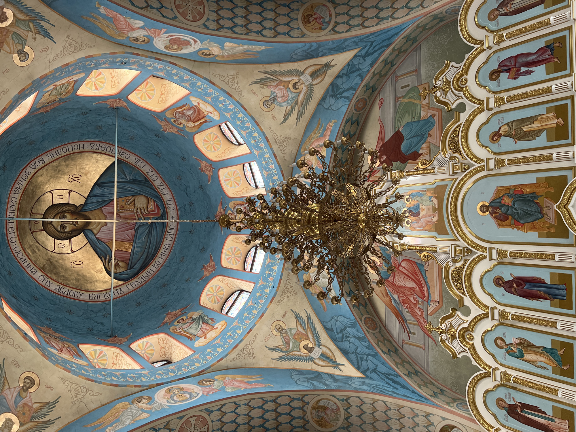 The image captures an interior view of a domed ceiling adorned with religious frescoes, specifically within a church in Kargopolye, Russia. Dominating the scene is a large, ornate chandelier crafted from gold-toned metal, suspended from the center of the dome. The dome itself is decorated with a series of religious paintings, primarily depicting biblical figures and scenes. The frescoes feature a palette of blues, reds, golds, and creams, and are framed by ornate architectural details. The presence of windows suggests natural light filtering into the space, enhancing the vivid colors of the artwork. In addition to the frescoes on the dome, the ceiling is further adorned with rows of rectangular panels depicting individual figures, each framed by elaborate gold-toned designs. The scene exudes an atmosphere of religious significance and artistic richness, characteristic of traditional Russian Orthodox churches.