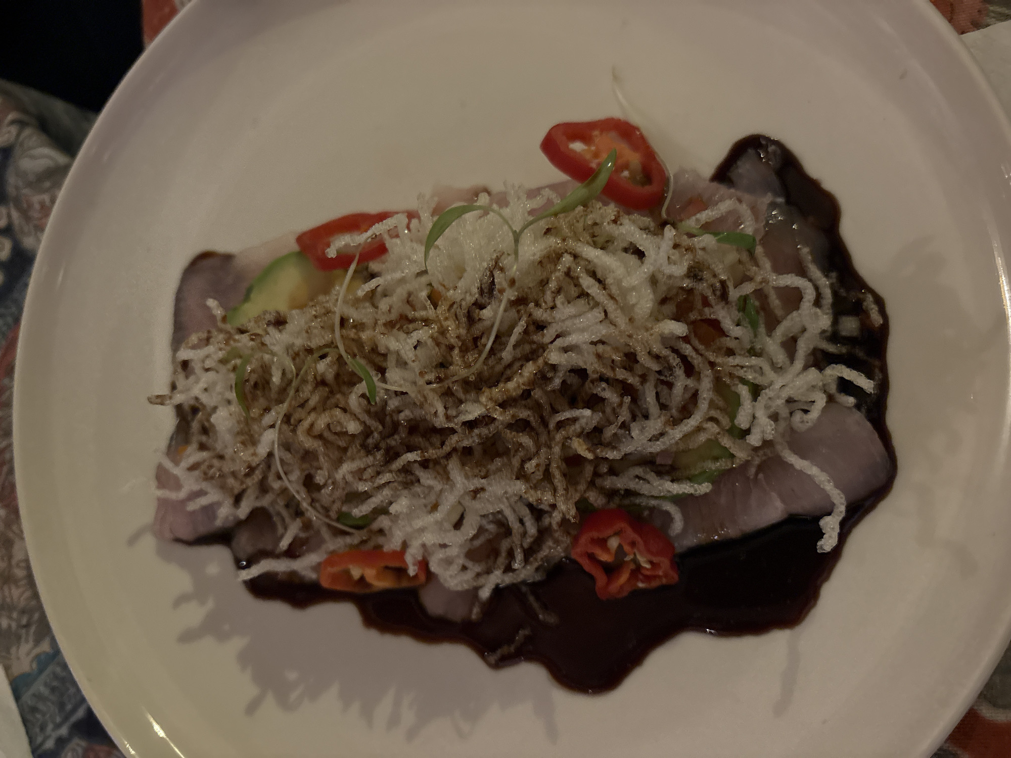 The image depicts a dish on a white plate. The dish consists of thinly sliced meat, possibly tuna or another firm-fleshed fish, arranged in a rectangular shape and drizzled with a dark sauce, likely a soy-based reduction. The meat is topped with shredded fried rice noodles, some thin slices of avocado, and thinly sliced red peppers. Some small green sprouts are also present. The plate appears to be placed on a tablecloth with a floral or paisley pattern. Given the dish presentation and ingredients, it is plausible this image was taken in Miraflores, Peru, known for its upscale dining and fusion cuisine restaurants.