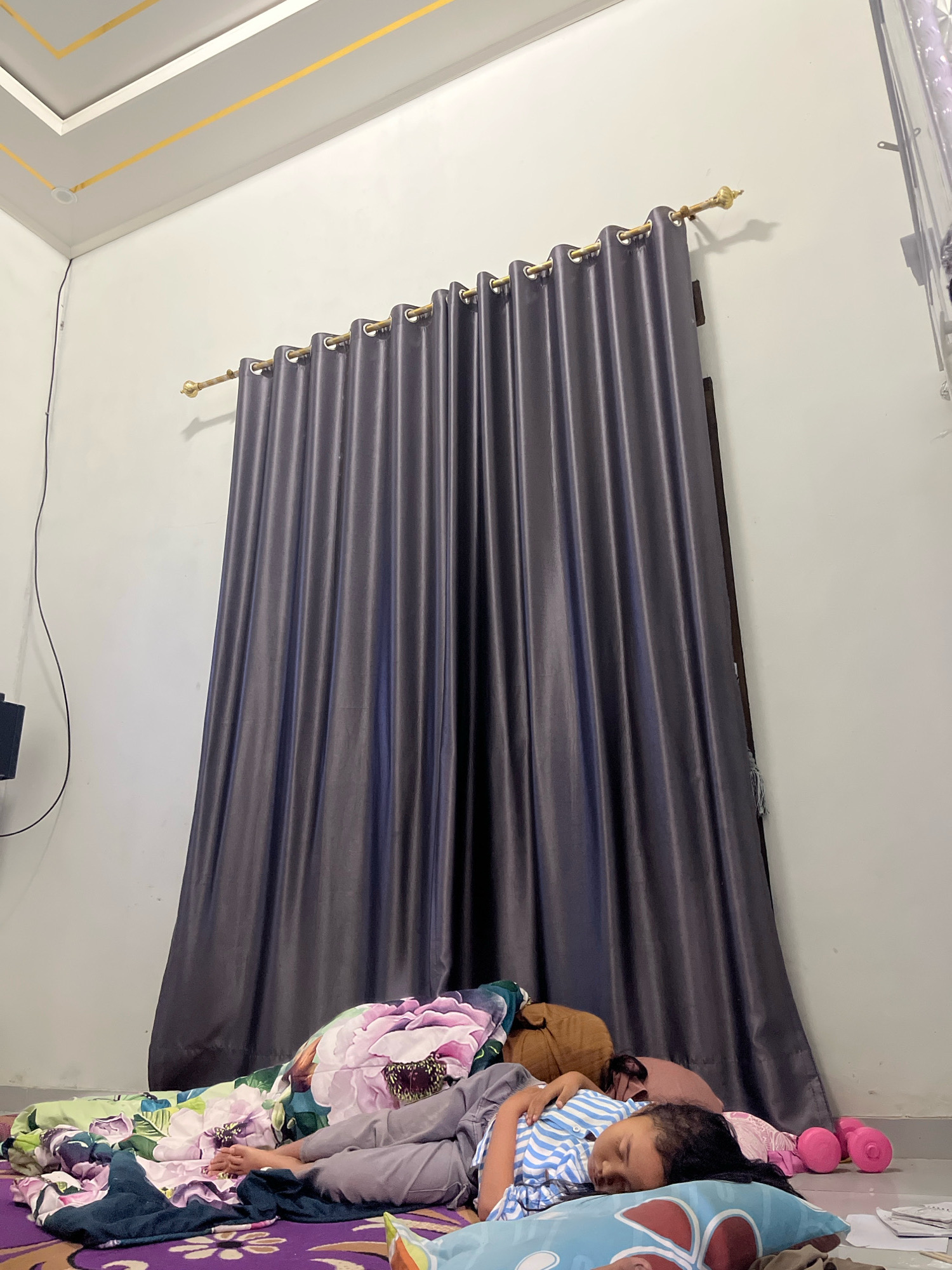 In a bedroom in Kebumen, Indonesia, a young girl is sleeping on the floor. She is lying on her side, on a pillow, with her face turned towards the camera. She is wearing a blue and white striped shirt and grey pants. Her legs are bent and her feet are visible. She is covered by a blanket with a floral pattern.
The room has white walls and a grey curtain hangs behind the girl. The curtain is drawn closed, obscuring any view of the outside. There are several pillows on the floor around the girl, and a brown stuffed animal is visible near her head. Pink dumbbells are also on the floor to the right of the girl. The ceiling is white with gold trim. The floor appears to be tiled. The overall impression is one of a child sleeping soundly in a comfortable, albeit slightly cluttered, bedroom.