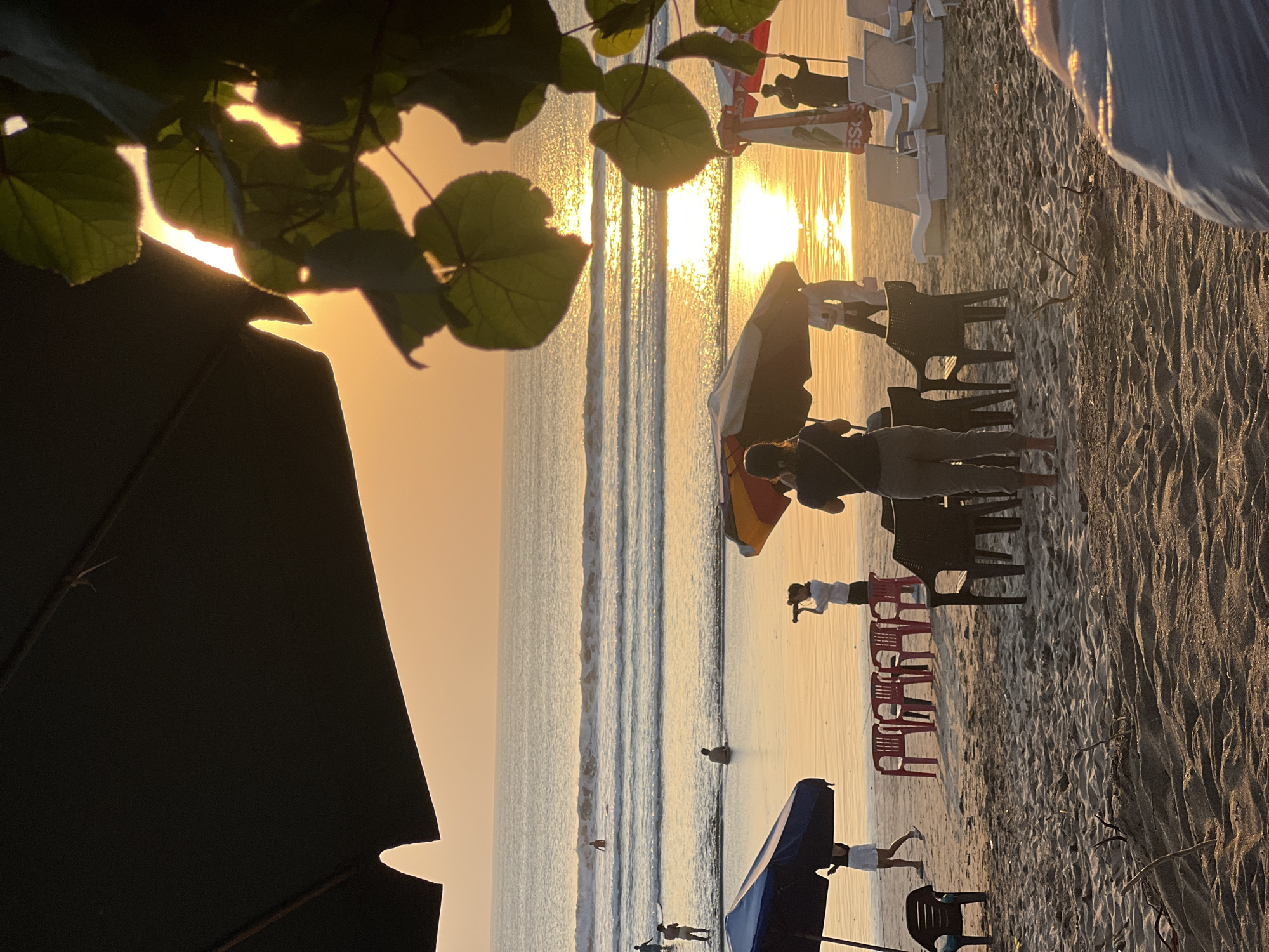 The image depicts a tranquil sunset scene at Kuta Beach, Bali, Indonesia. The sky is ablaze with a warm orange hue, casting a golden glow over the ocean and the sandy shore. Gentle waves roll onto the beach, creating a soothing rhythm. Several people are visible enjoying the sunset: a woman stands with her back to the camera, looking out at the ocean; another woman is taking a photo with a telephoto lens; and further out in the water, individuals are seen swimming and surfing. Beach chairs and umbrellas are set up, suggesting a relaxed atmosphere. The foreground is dominated by sand dunes and a few pieces of driftwood, adding to the natural beauty of the scene.