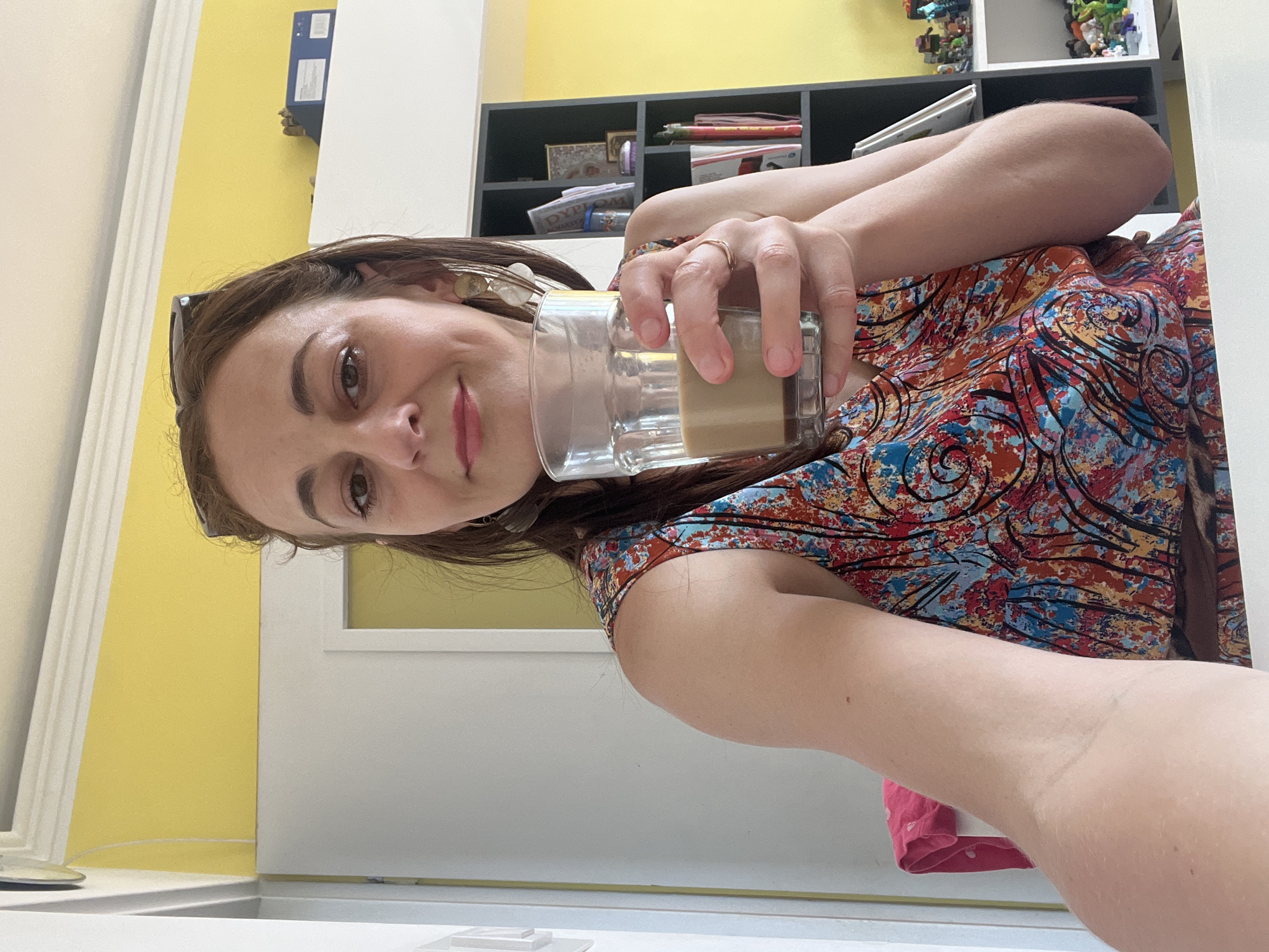 A woman stands in an indoor setting, holding a glass of coffee with her right hand. She is wearing a colorful sleeveless dress and has a pair of sunglasses perched on her head. Her left arm is extended outwards, suggesting she is taking a selfie. Behind her, a bookshelf is visible, filled with books and decorative items. The background features a bright yellow wall. The location is described as Unknown, Unknown.