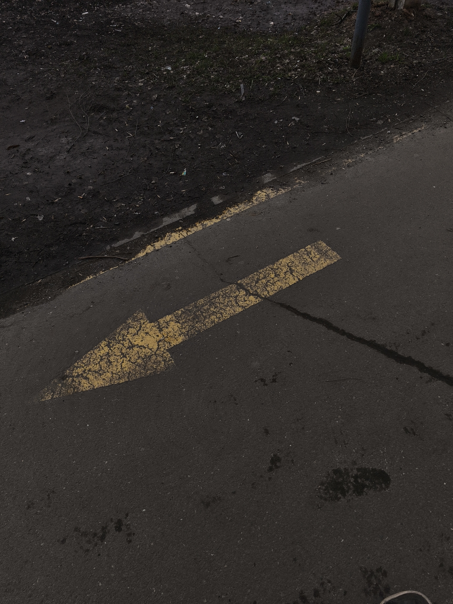 The image captures an outdoor ground-level scene in Izhevsk, Russia, depicting a section of dark asphalt next to a muddy, unkempt strip of ground. A prominent, worn yellow arrow is painted on the asphalt, pointing towards the upper left portion of the frame. The yellow paint shows signs of cracking and fading. A thinner yellow line runs along the edge of the asphalt, separating it from the adjacent dirt area.
The muddy ground is dark brown to black, appearing wet and uneven, with scattered small twigs, dried leaves, and what appears to be minor debris or litter. Patches of sparse, dull green and brown vegetation are visible within the dirt. In the upper right corner, a dark, vertical pole is partially obscured by the frame, rising from the grassy edge.
The asphalt surface itself is a dark gray, textured, and marked with various dark, wet-looking stains and streaks, particularly in the lower right section, suggesting recent dampness or rain. The lighting is diffuse and even, indicating an overcast sky or low light conditions. No specific activity is occurring, and there are no people or animals visible. The only visible marking is the directional yellow arrow. The scene conveys an ordinary, somewhat neglected outdoor pathway.