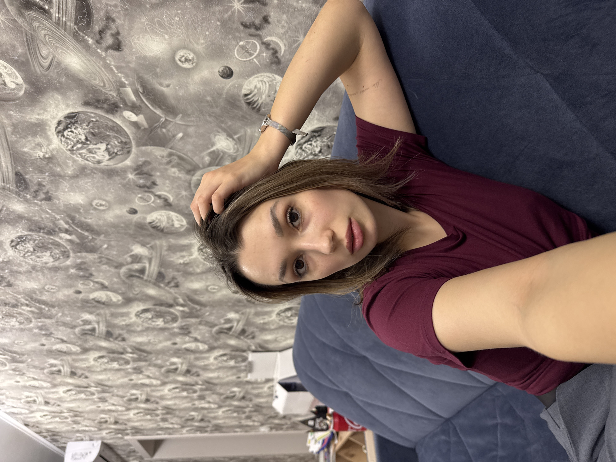 A woman is reclining on a blue couch, taking a selfie. She is wearing a maroon shirt. Her right arm is bent and held above her head, with her hand touching her hair. She has a watch on her wrist. The background is a wall with a cosmic pattern.