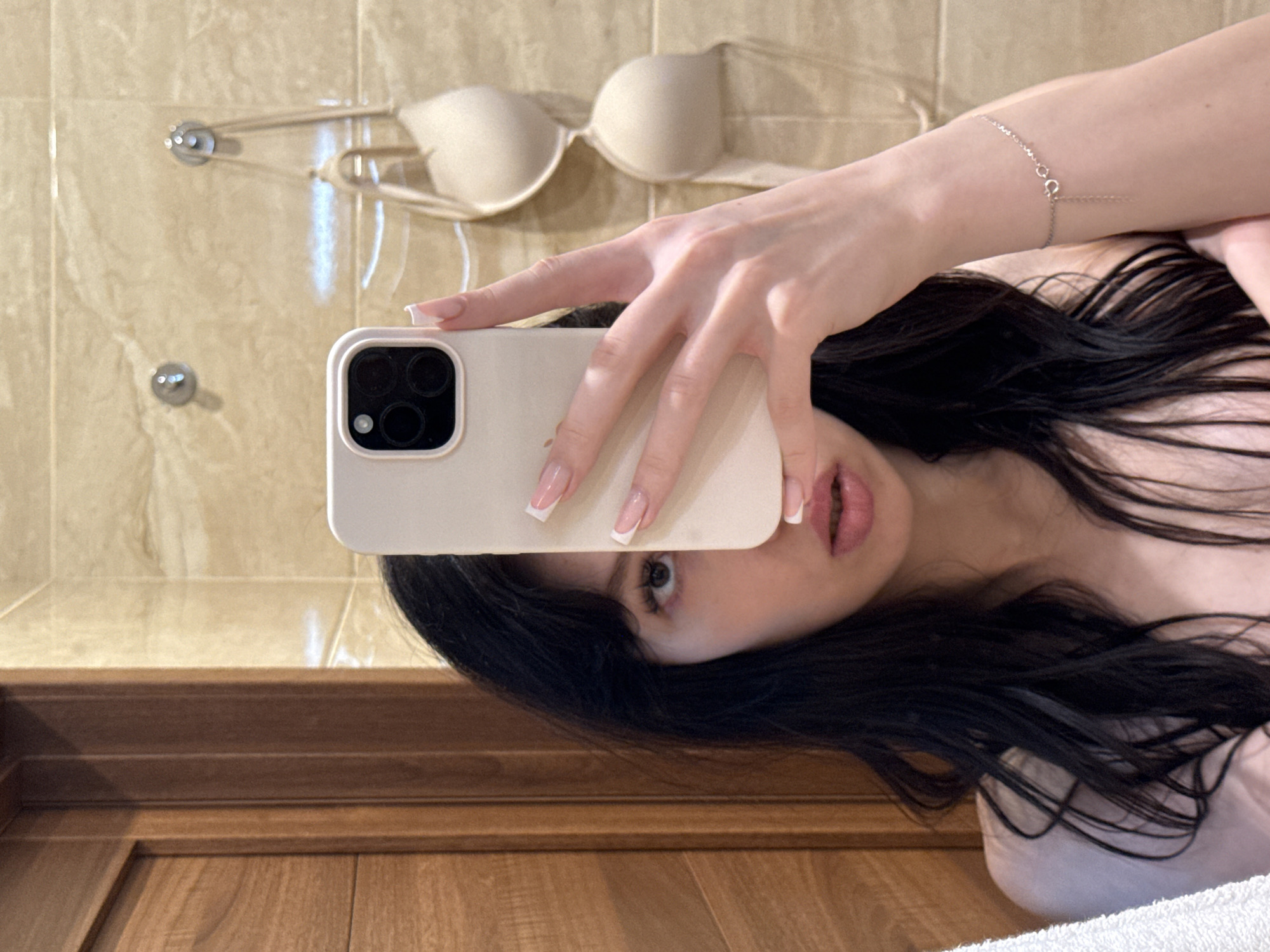 A young woman with long, dark hair takes a mirror selfie in what appears to be a bathroom. She is holding a light-colored smartphone in her right hand, obscuring the left side of her face. Her left eye is visible, and she has on a light-colored top that is mostly out of frame. Her nails are painted white. In the background, a wooden door is visible on the left, and tiled walls with two hooks holding a beige bra are on the right. A silver bracelet is on her left wrist.