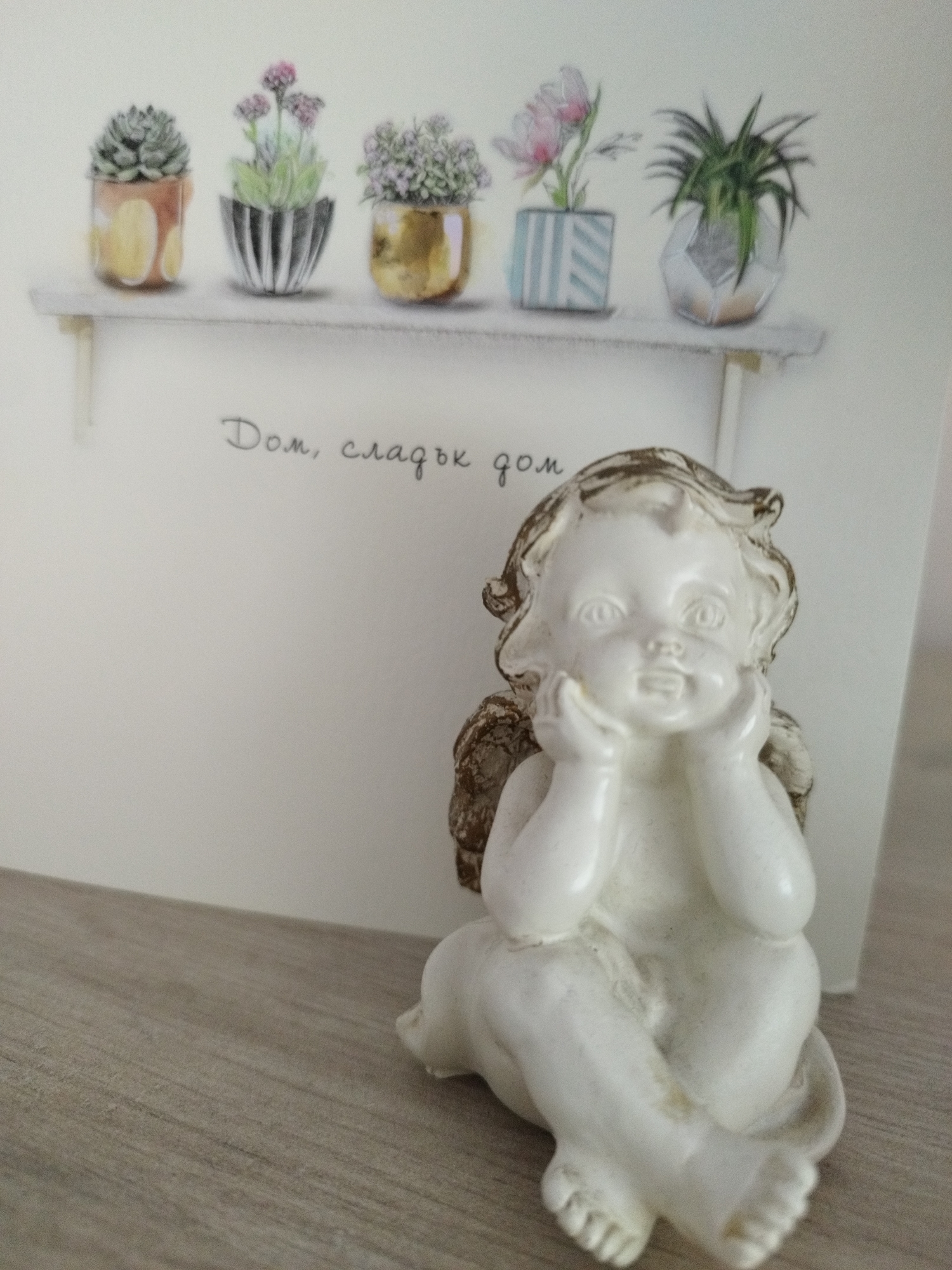A close-up shot shows a small, white angel figurine sitting on a light grey, wood-grain surface. Behind the figurine is a card. The card shows five potted plants on a shelf, along with some Bulgarian text, "Дом, сладък дом," which translates to "Home, sweet home." The location of the scene is not discernible from the given information.