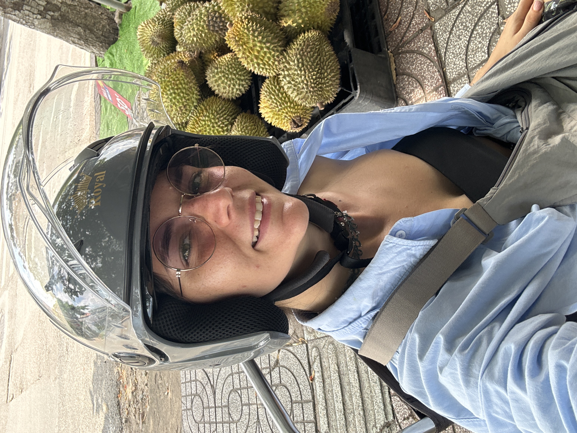 A close-up outdoor shot depicts a young adult female seated, wearing a grey open-face motorcycle helmet with a clear visor. The helmet features a gold "Royal" brand logo with a crown icon on the front. She wears round-frame glasses and smiles, revealing her teeth. Underneath the helmet, a black chin strap is secured, and a dark, multi-charmed neck strap is visible. Her attire consists of a light blue button-up shirt, unbuttoned to expose a black top or bra, and a grey shoulder strap from a bag across her chest. Her right hand is visible in the lower right, resting on what appears to be a bag or her lap.
To the right and slightly behind the woman, a significant pile of durians with green-yellow thorny exteriors is displayed in black plastic crates, resting on a green mat or tarp. A tree trunk is visible behind the durians. Reflections in the helmet's visor reveal a partial red sign with white text, possibly indicating a price or promotion. The background consists of a grey patterned tiled sidewalk in the foreground and a textured asphalt road further back. A silver metal bar, potentially from a scooter or motorcycle, is visible on the left side. The scene is situated in Phu Thuy Ward, Vietnam, suggesting a street market or roadside vendor setting.