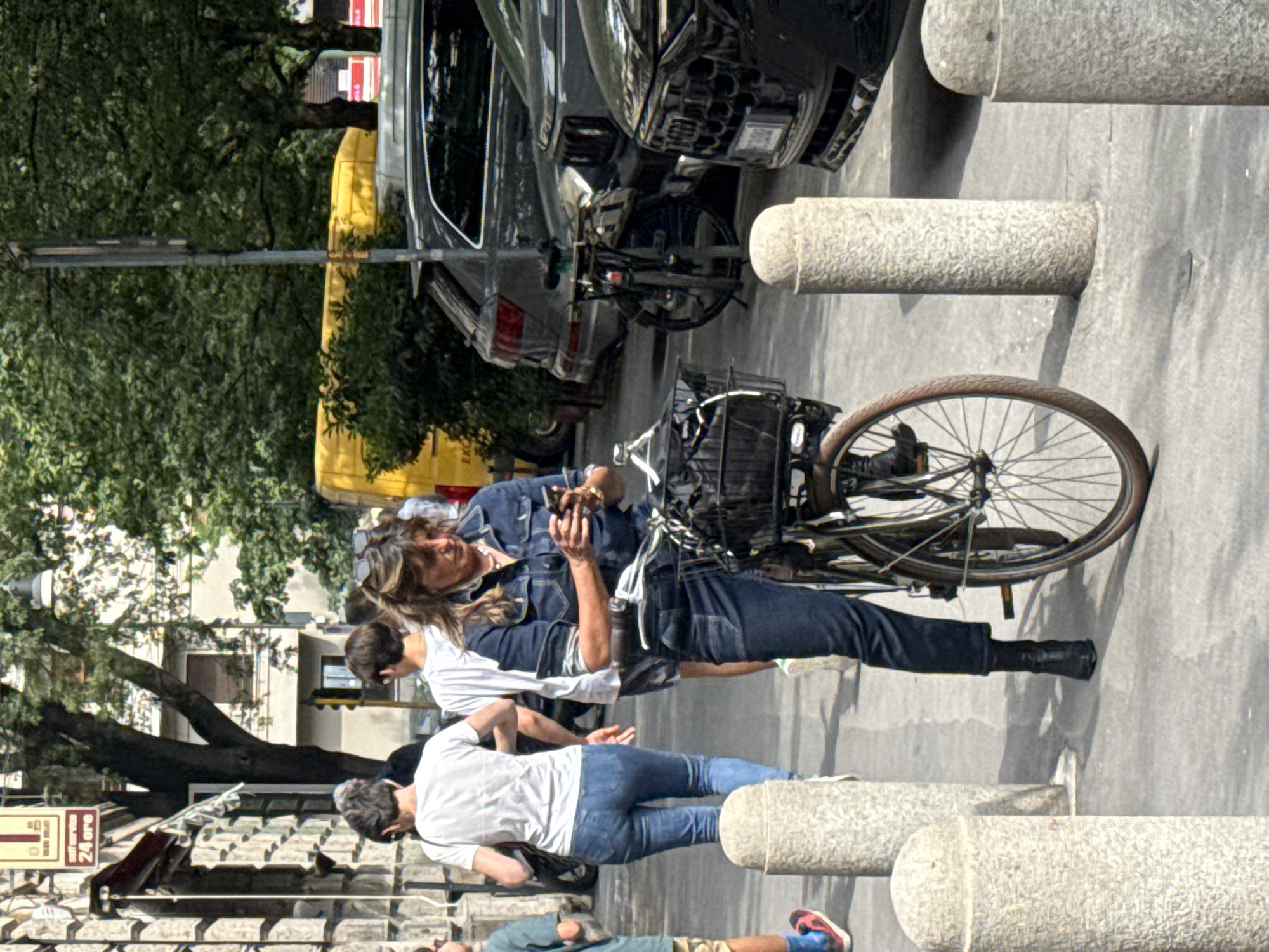 The image captures a sunlit street scene in what appears to be Milan, Italy. The setting is an urban environment featuring a sidewalk lined with stone bollards, adjacent to a street with parked vehicles and trees.
In the foreground, a woman with long, wavy brown hair, dressed in a denim jacket and blue jeans, is standing beside a bicycle. She is holding and looking at a smartphone, seemingly engrossed in its content. Her bicycle has a basket mounted on the handlebars. Behind her, another person in a white t-shirt and jeans is partially visible, talking to someone further back. A third individual, also in a white t-shirt and jeans, is seen looking downwards. In the lower left corner of the frame, parts of a fourth person wearing a blue shirt, shorts, and red sneakers are visible.
The background features parked cars, including a black SUV and a yellow van. Lush green trees with dense foliage cast shadows, indicating bright daylight, likely midday or early afternoon. Buildings with classic European architecture are visible through the trees on the left side of the frame.
The activity depicted is a casual moment on a city street, with individuals either pausing or moving through the area. The woman with the bicycle appears to be taking a break to use her phone.
Notable details include the textured, grey stone bollards on the sidewalk. On a building facade in the background, a red and white sign is visible with the text "24 ore" (Italian for "24 hours"), confirming the location as Italy and likely Milan as specified. The weather is clear and sunny.