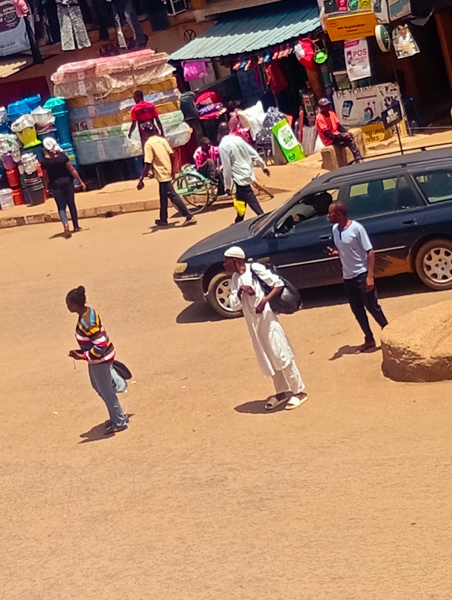 The image shows a street scene in an urban area, possibly in a city in an unspecified location. The environment is sunlit, suggesting a daytime setting. In the foreground, there is a light brown or sandy area. Two individuals are walking across it, one in a colorful striped sweater and jeans, and another in traditional white attire with a cap, carrying a black bag. A car is parked nearby, with another man in a blue shirt walking beside it. In the background, various street vendors are displaying goods such as mattresses, buckets, and clothing items under awnings. Signs are visible, including one for "POS" services and another advertising SIM cards. People are standing and sitting around the vendors, engaged in potential commercial activities. A wheelchair is visible near the vendors. The scene depicts a busy street with pedestrian and vehicular traffic.