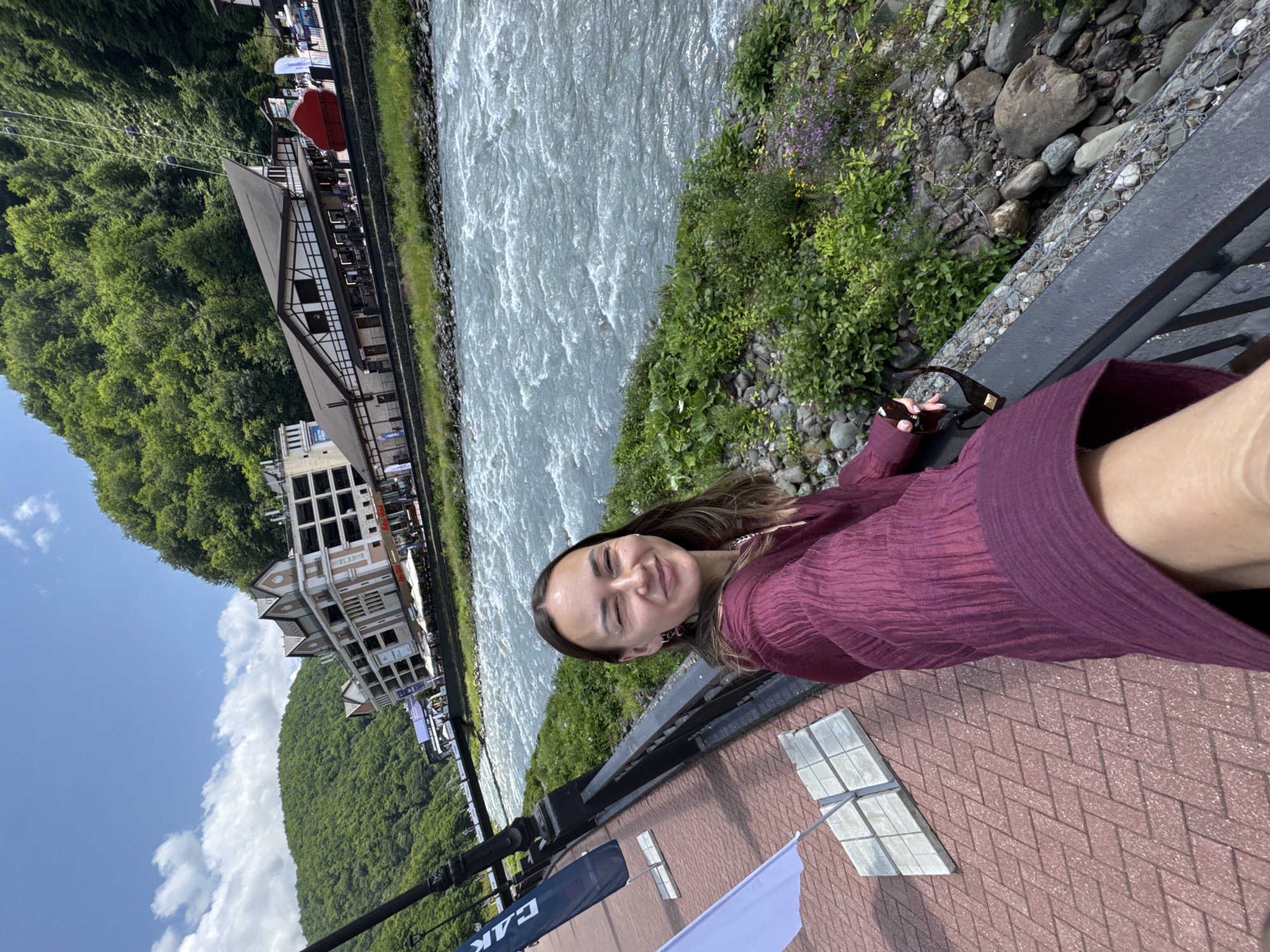 A young woman with brown hair and brown eyes is taking a selfie on a bridge with a river and mountains in the background. The woman is wearing a purple shirt and sunglasses. She is smiling at the camera. The river is wide and fast-moving. The mountains are covered in trees. The city of Estosadok, Russia is visible in the background. The sky is blue with some clouds. There are flags on the left side of the image.