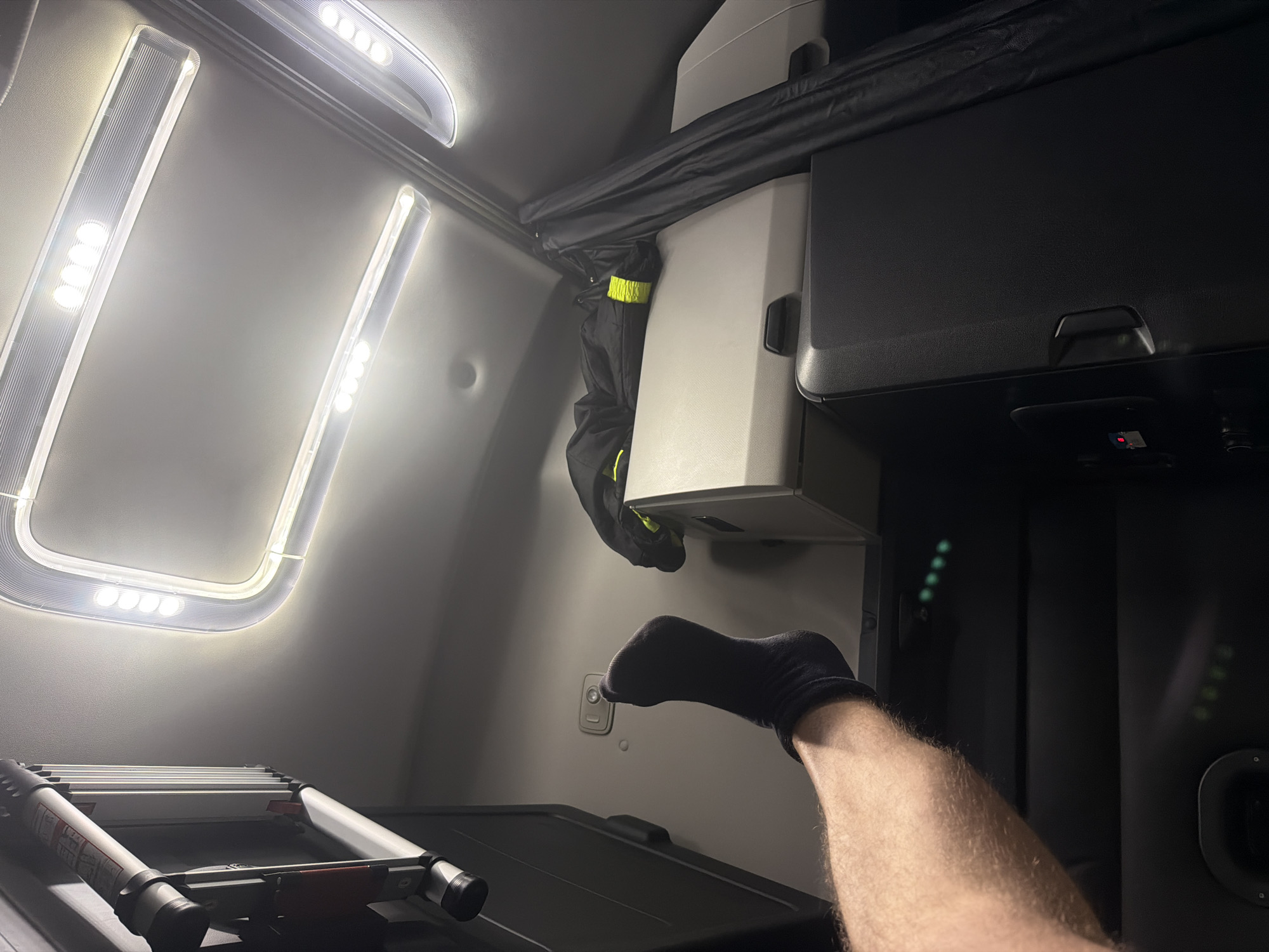 A person's leg, wearing a dark sock, is propped up on a surface inside a vehicle. The leg is positioned from the lower thigh to the foot. Above the leg, the interior of what appears to be a truck cab is visible, featuring ceiling lights and a folded garment with reflective yellow detailing draped over a storage unit. A black curtain hangs to the right. In the background, part of an extendable ladder is mounted vertically on the left side. The lighting in the scene is artificial, with bright overhead lights and some green indicator lights visible lower down. The overall environment suggests the interior of a commercial vehicle, possibly a truck, in a setting that is not specifically identifiable as New Britain, United States of America.