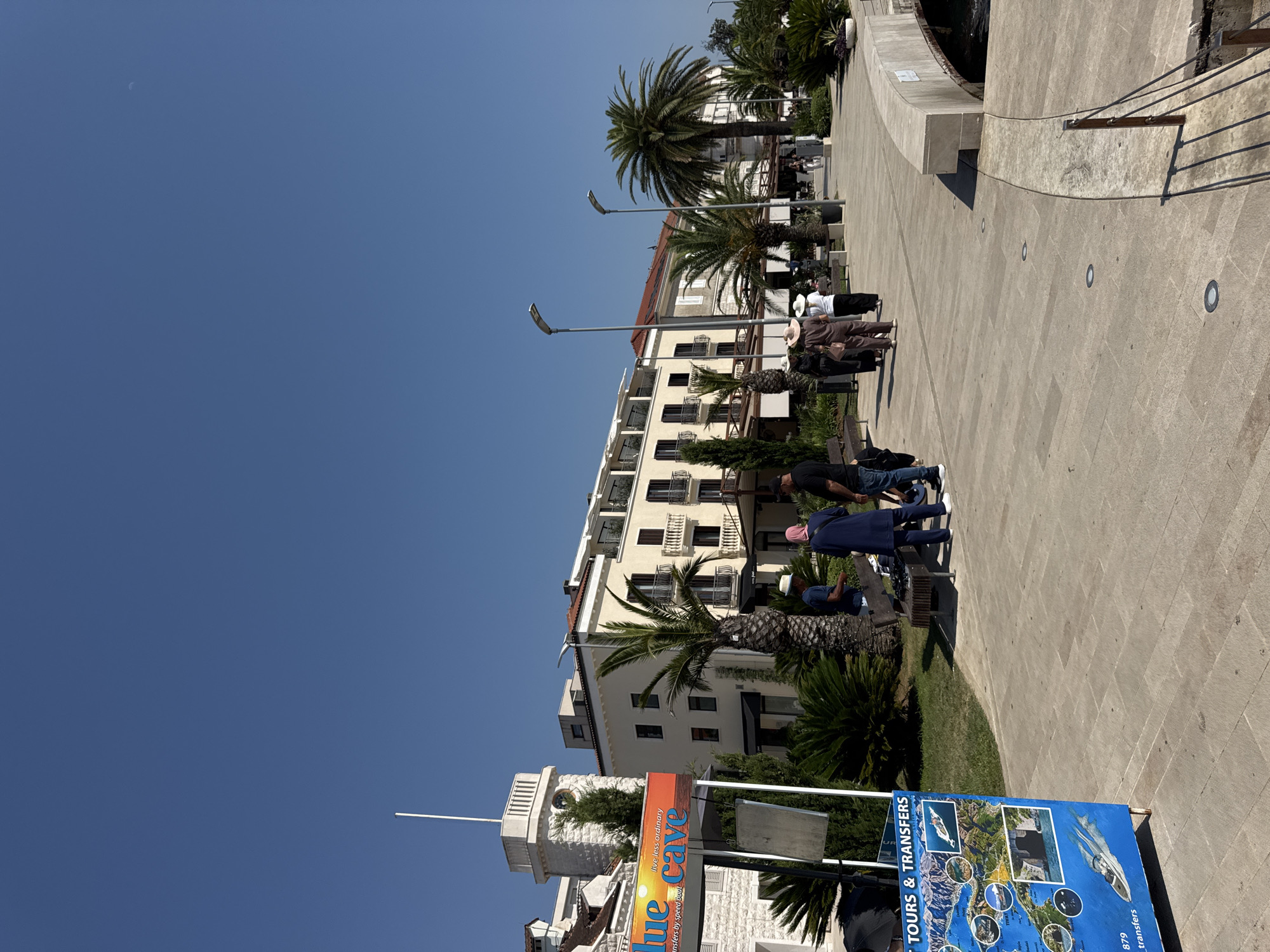 The image depicts a sunny day in Tivat, Montenegro, likely along the waterfront promenade. In the foreground, a blue sign advertises "Blue Cave Tours & Transfers," featuring images of boat trips and destinations. Several people are visible, some gathered around the sign, including a person in a blue headscarf and another in a straw hat. Further down the promenade, more individuals walk or stand in small groups, with a focus on the architecture of the buildings lining the street.
The buildings are a mix of styles, with a prominent cream-colored building featuring balconies and a red-tiled roof. To the left, a white stone tower with a small window and a flagpole stands out. Lush greenery, including palm trees and other ornamental plants, dots the landscape, creating a Mediterranean ambiance. The walkway is paved with light-colored stone, and streetlights are interspersed along the path. A curved concrete bridge or embankment is visible on the right, with dark water beneath. The clear blue sky suggests fair weather.