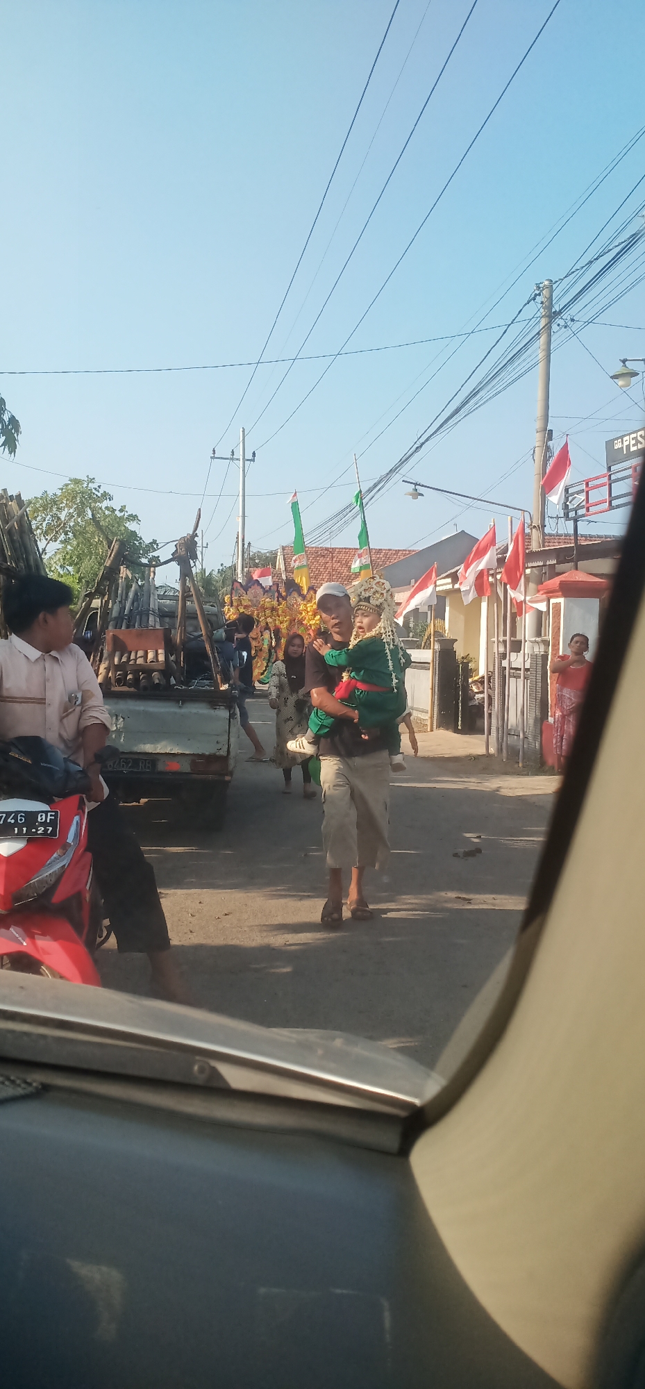 In Kalisalam, Indonesia, a procession is underway on a paved road under a clear blue sky, with power lines crisscrossing overhead. A man is carrying a child dressed in traditional attire on his shoulders. They are walking past a pickup truck laden with bamboo poles. Several people are walking alongside, and some are in the vicinity of a building adorned with Indonesian flags. A red motorcycle is parked on the left side of the road, and part of a car's interior is visible in the foreground, suggesting the photo was taken from inside a vehicle.