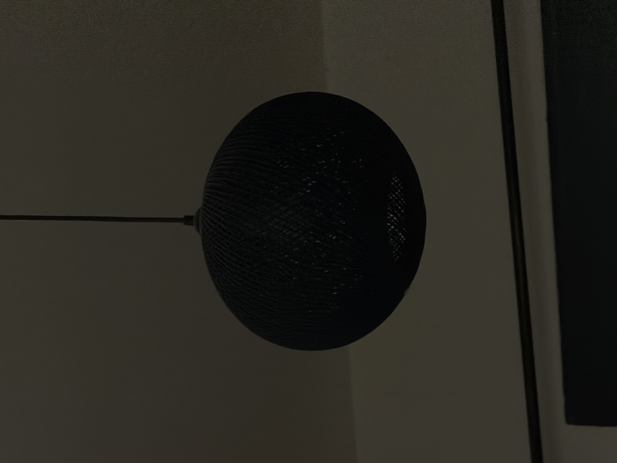 The image features a spherical lampshade constructed from woven material, likely placed indoors against a neutral-toned wall. The lampshade is black and appears to be suspended by a thin, dark rod. A framed picture, partially visible on the right, has a black border and a dark or black content area. The overall lighting in the scene is dim, and the setting is an interior space. There's no indication of the location being in Podgorica, Montenegro, or anywhere else.